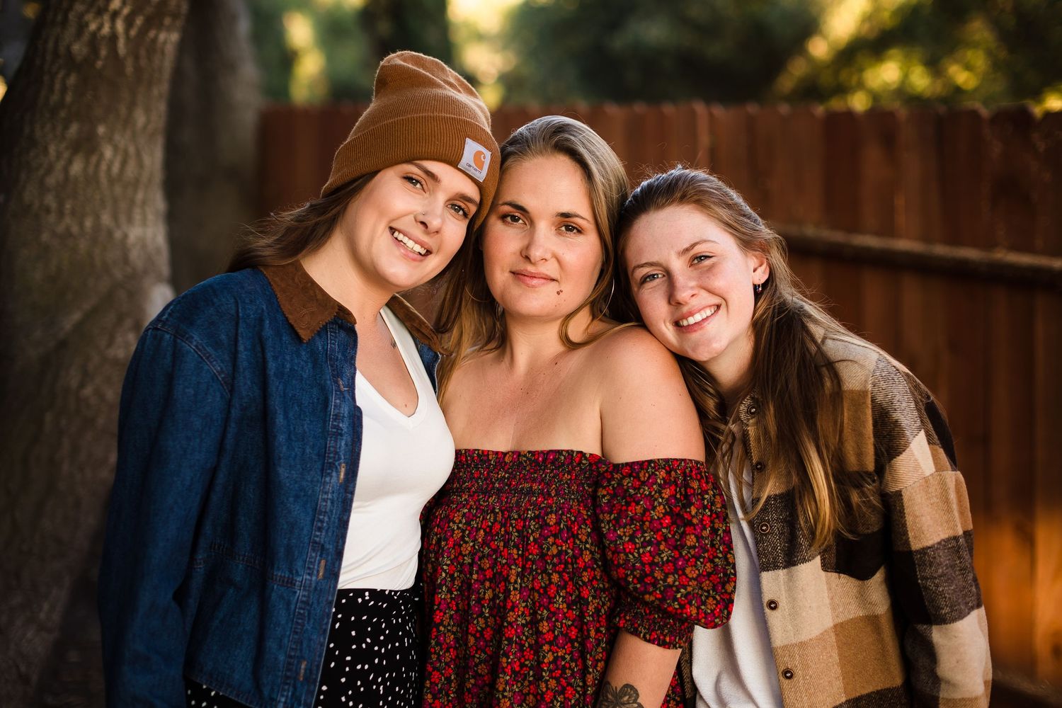 Three people smiling and embracing outdoors during golden hour with a wooden fence in the background.