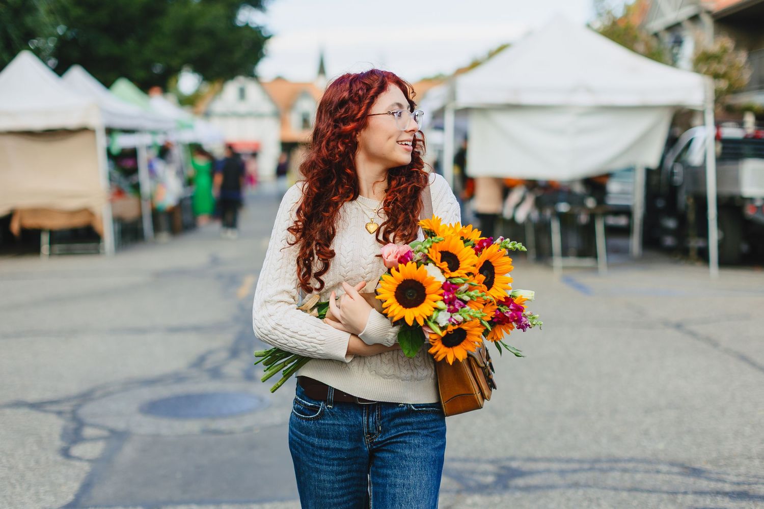 Red-haired woman holding colorful sunflower bouquet at outdoor farmers market, wearing glasses and cream sweater.