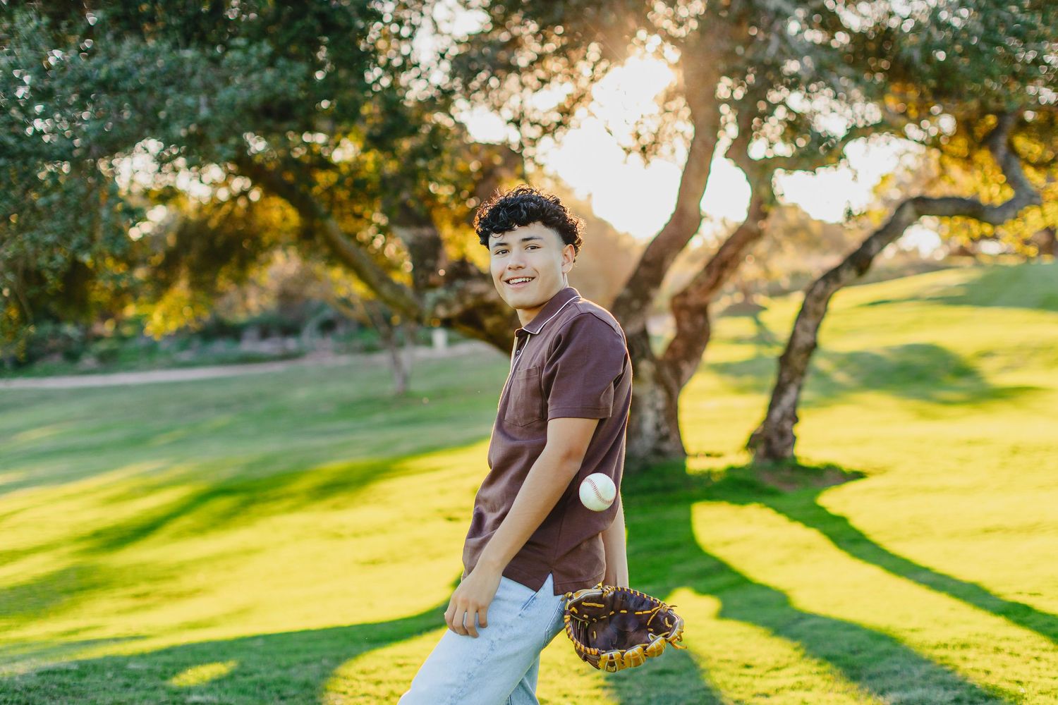 A smiling boy holds a basket of cherries in a sunlit park with lush green grass and trees in golden hour light.