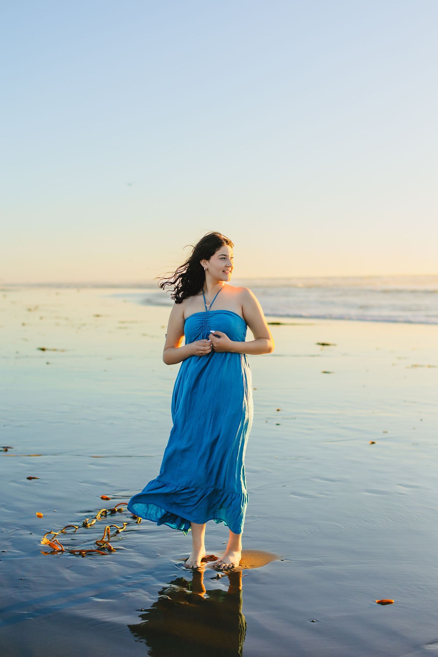 A smiling woman in a flowing blue strapless dress stands barefoot on a wet beach at sunset.