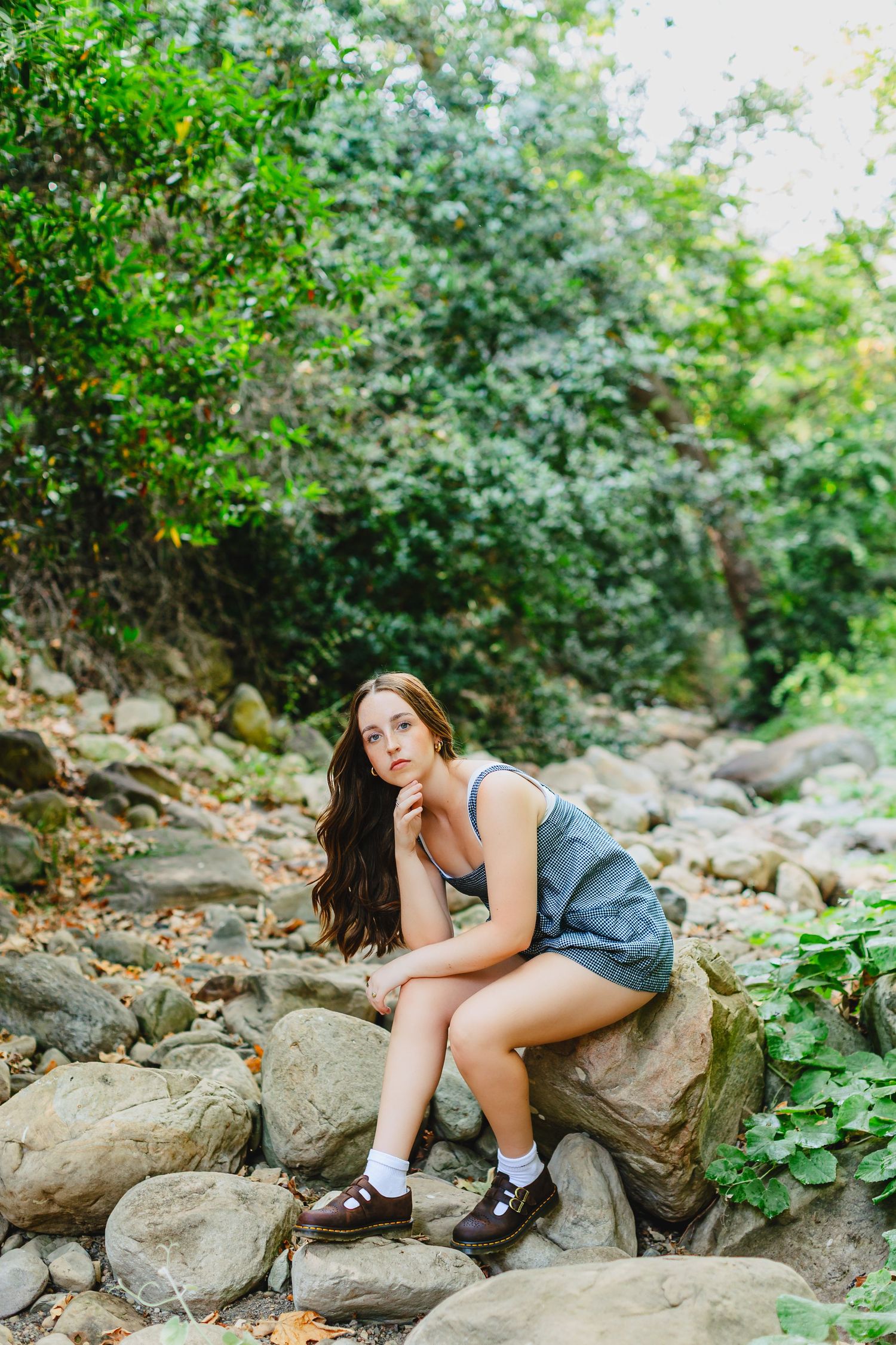 Young woman in gray romper sitting on rocks in a lush green forest setting, posing thoughtfully.