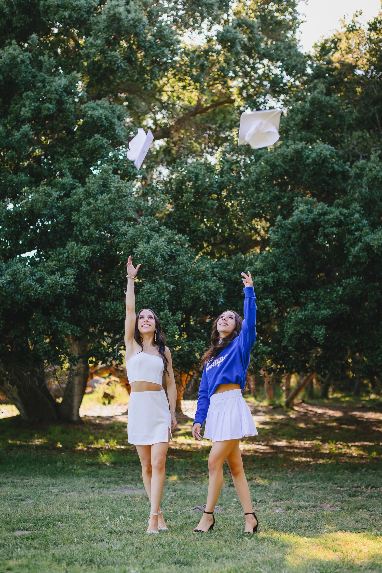 Two graduates toss their caps in celebration under green trees on a sunny day.