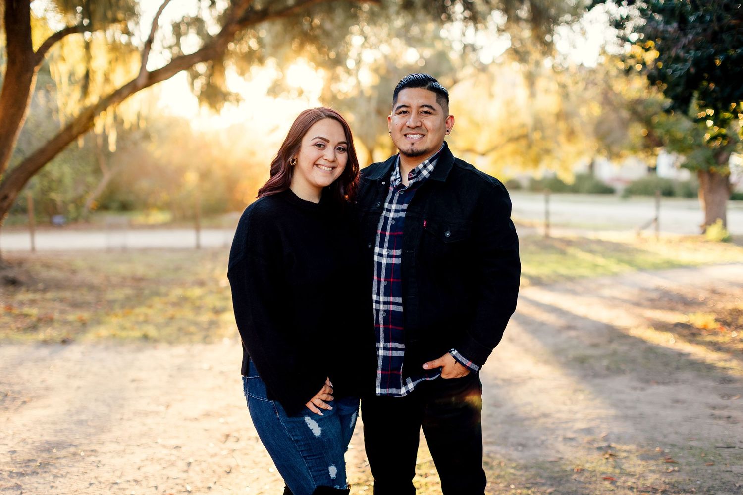 Couple in coordinating black and plaid outfits pose together during golden hour in an outdoor nature setting.