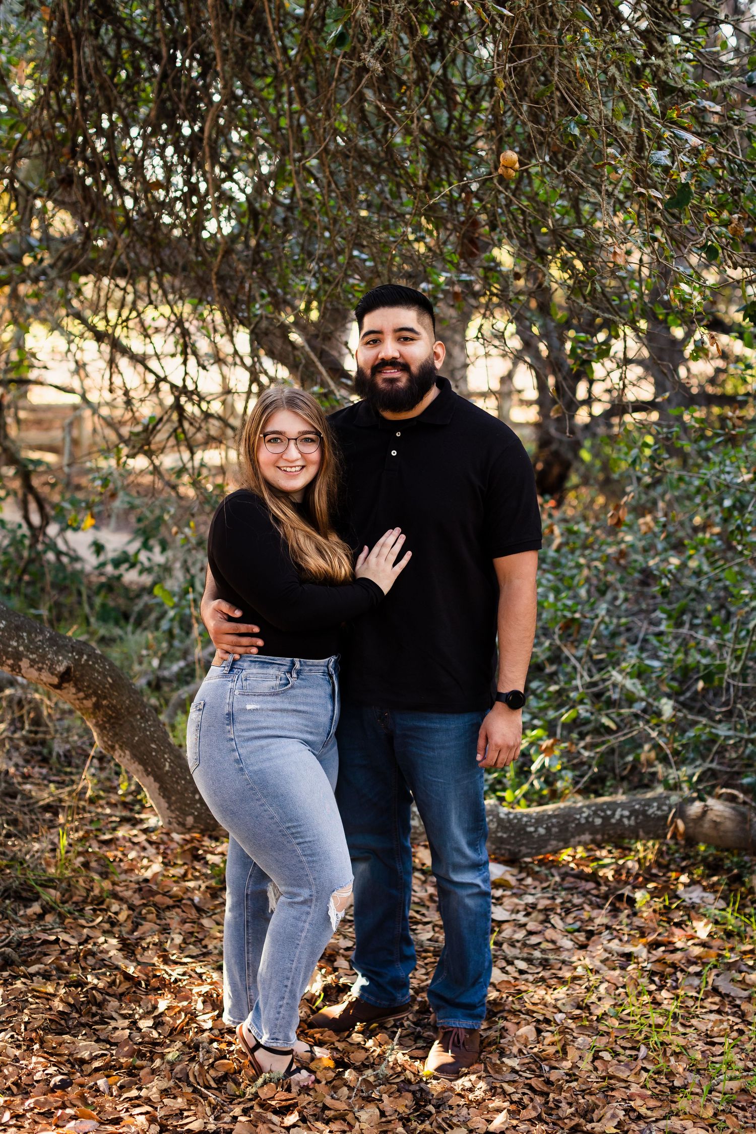 A couple in casual attire embraces while standing under a tree in a natural outdoor setting during autumn.