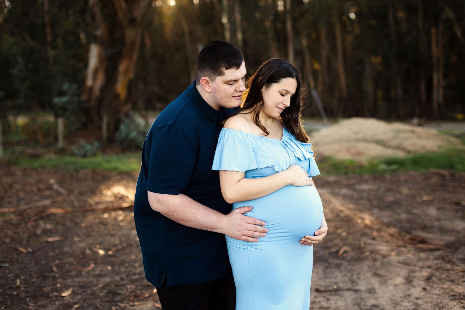 A couple embraces during a romantic outdoor maternity photoshoot at golden hour in a wooded setting.