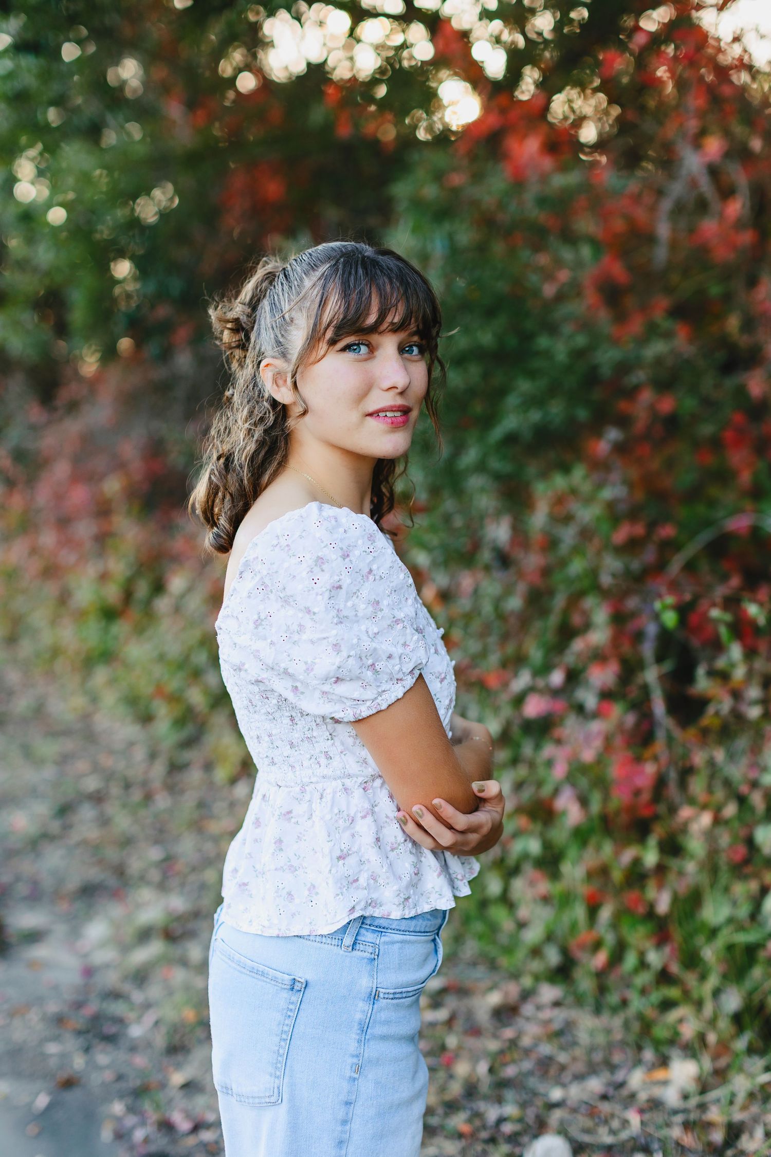 Young woman in white floral top and jeans poses outdoors among vibrant red and green autumn foliage.