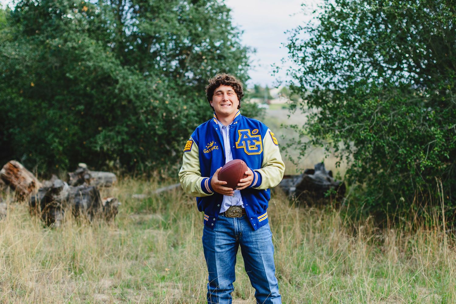 Young man in a blue varsity jacket holding a football, smiling outdoors in a grassy field with trees.
