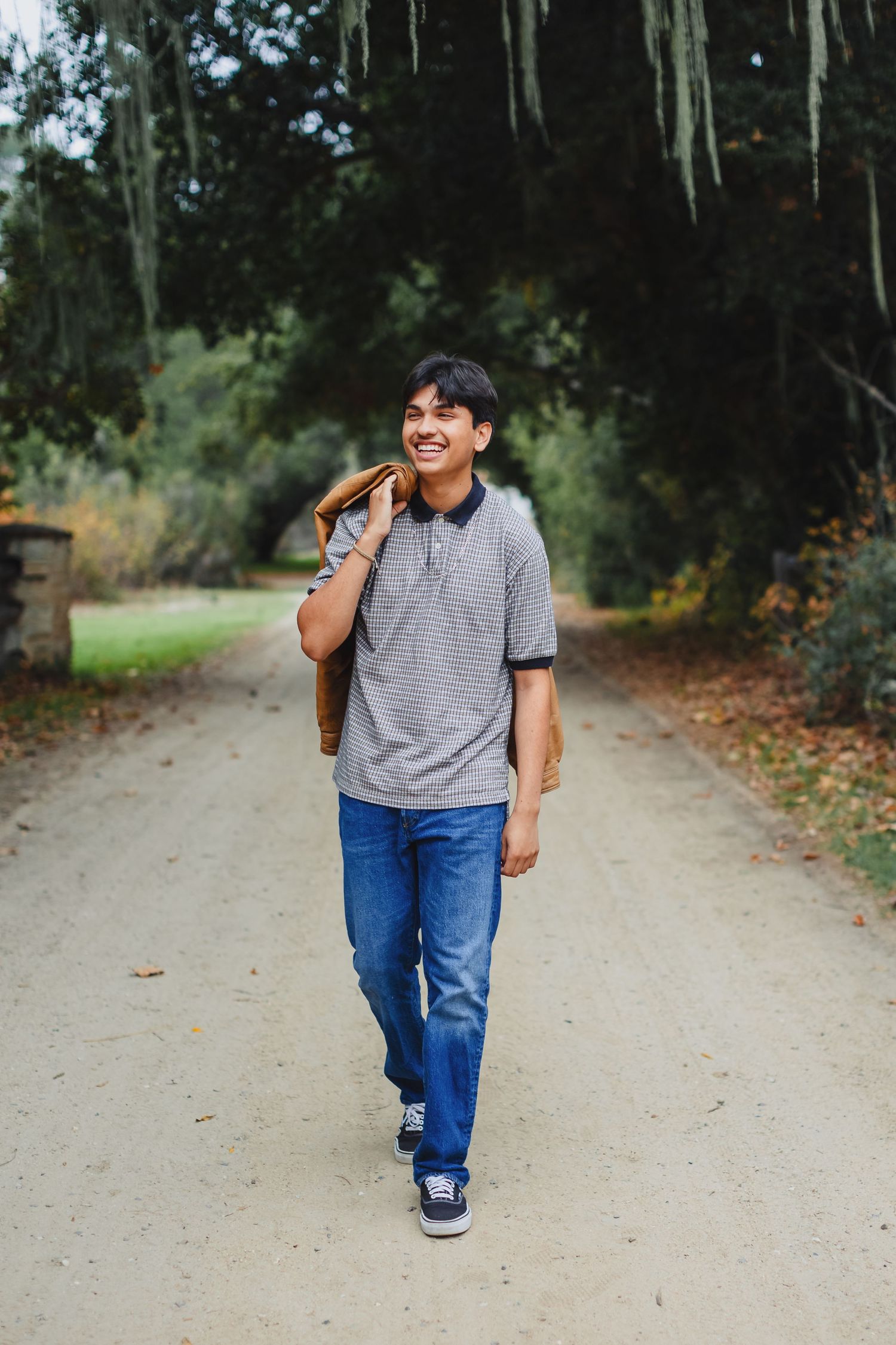 A person in a striped t-shirt and jeans walks smiling down a tree-lined path on a sunny fall day.