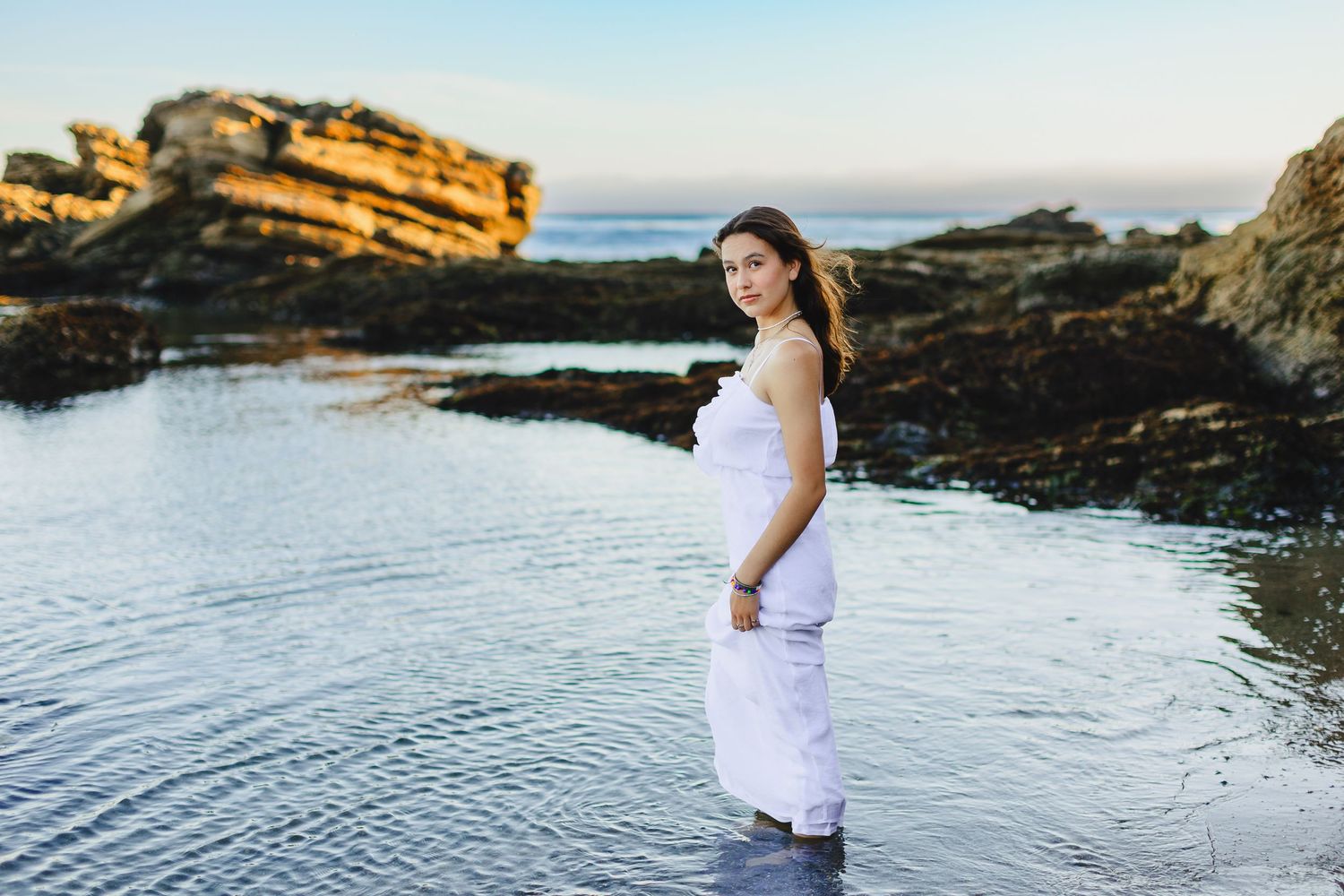 Pregnant woman in white dress stands in shallow coastal tide pools at golden hour.