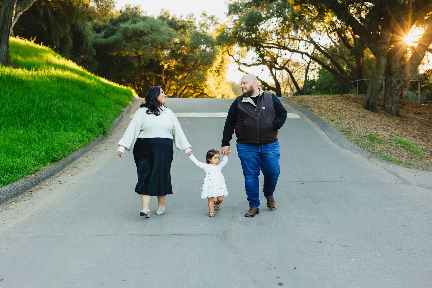 Family of three walking hand-in-hand down a sunlit park path at golden hour.