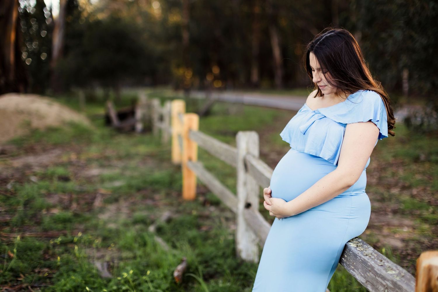 An expectant parent wearing a light blue dress poses in multiple photos at a rustic wooden ranch fence during sunset.