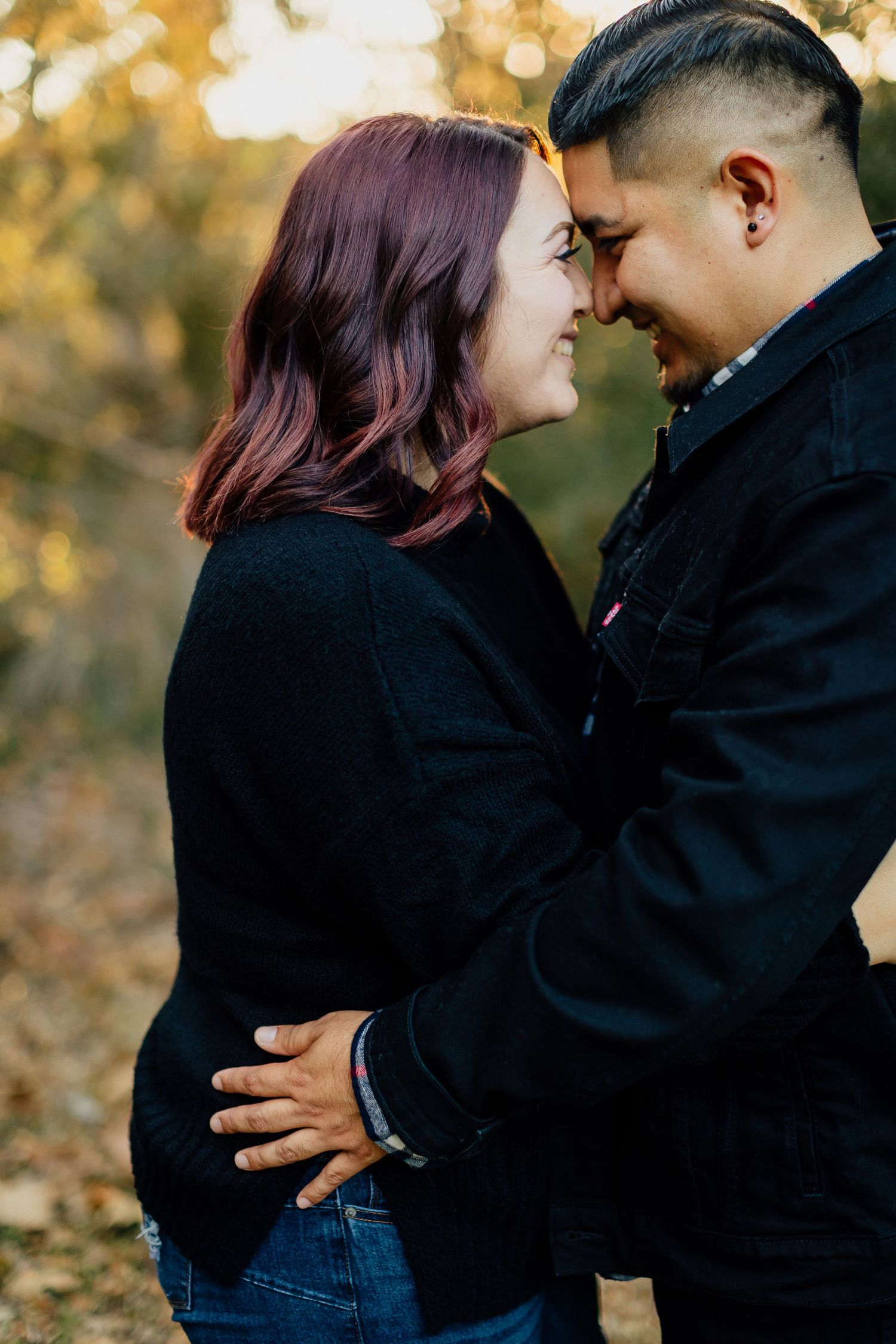 A couple in black sweaters embraces tenderly during an autumn outdoor photoshoot with golden bokeh lighting effects.