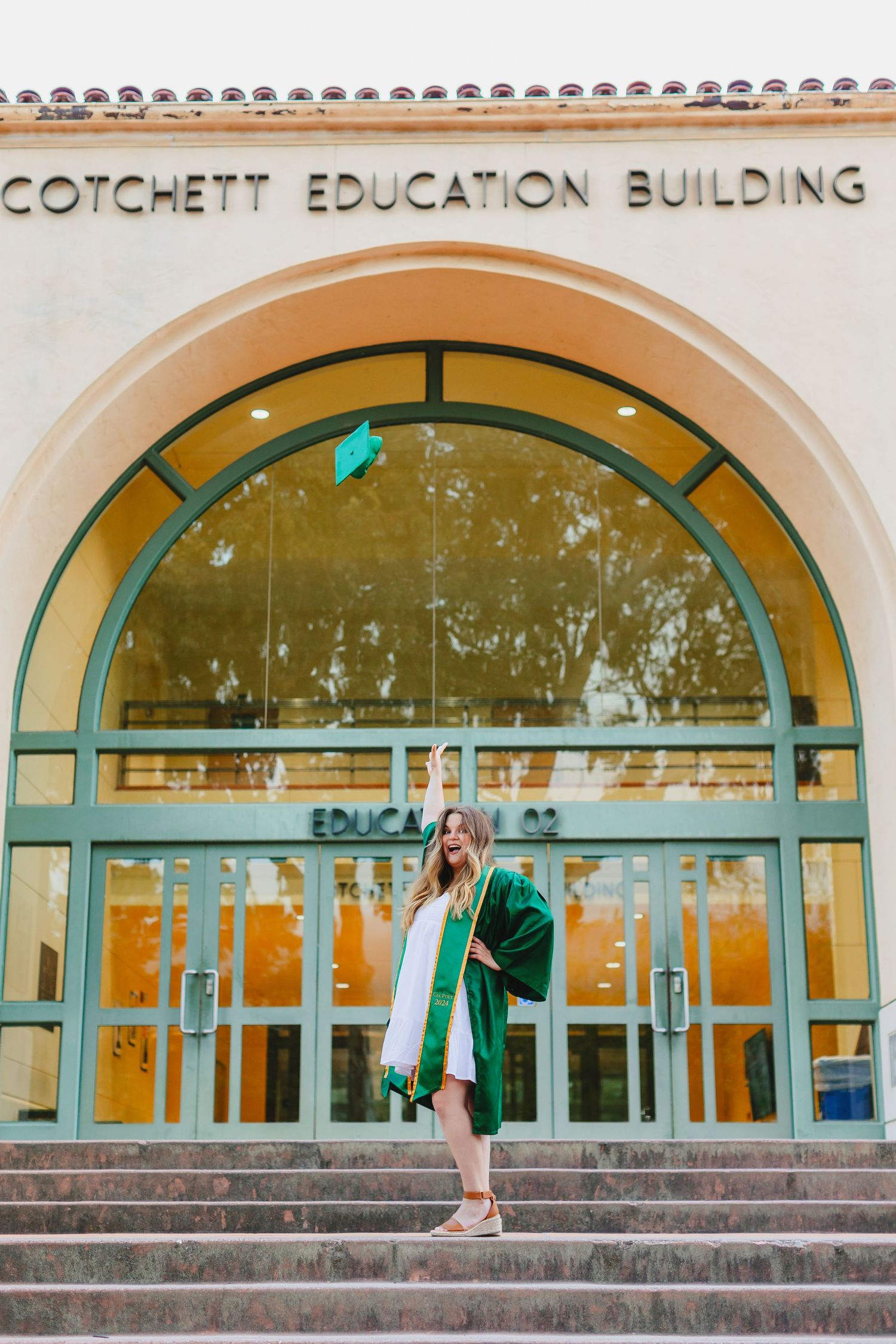 Graduate poses on steps of Cotchett Education Building in green graduation attire.