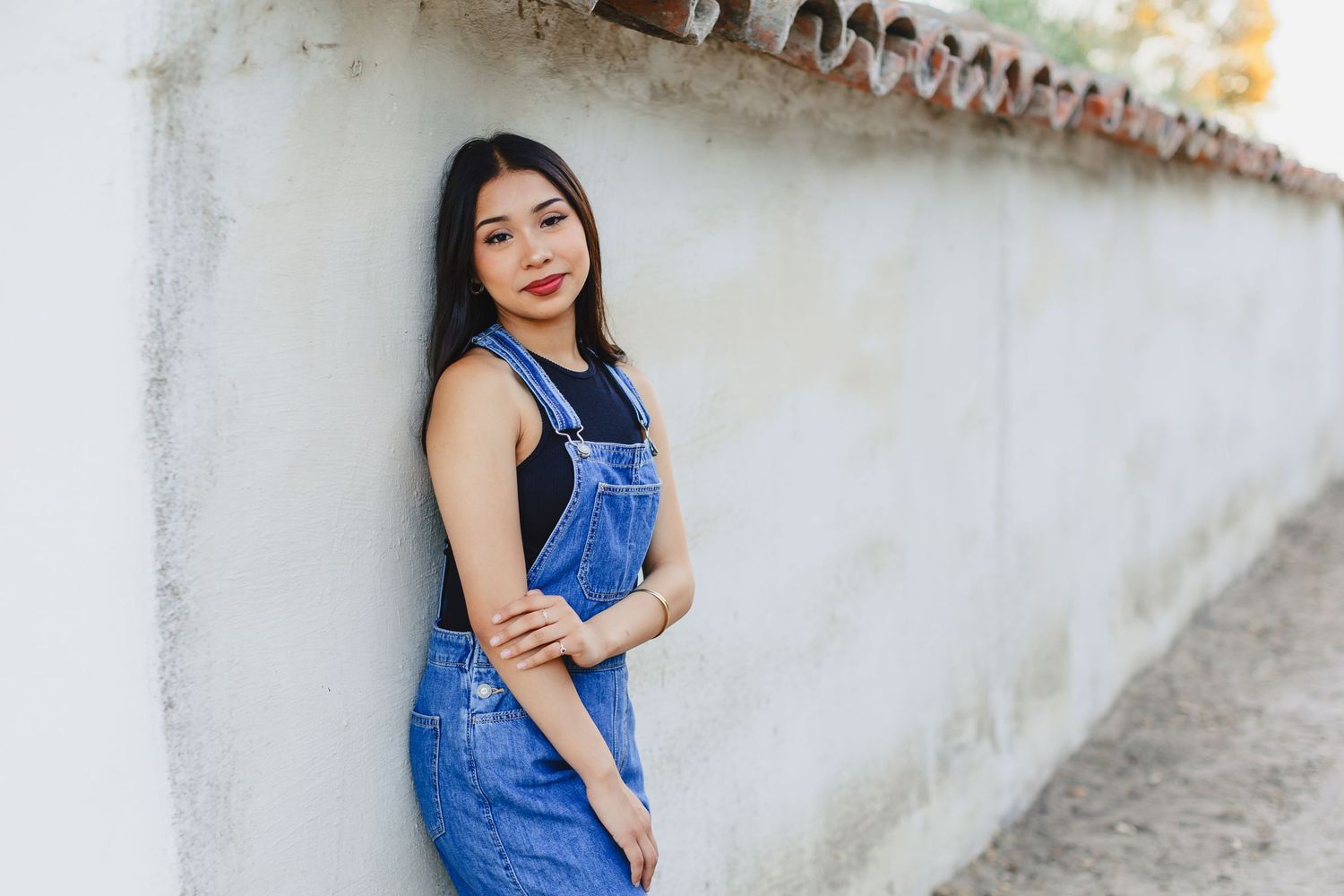 A woman in denim overalls stands with a confident pose against a white wall in casual summer style.