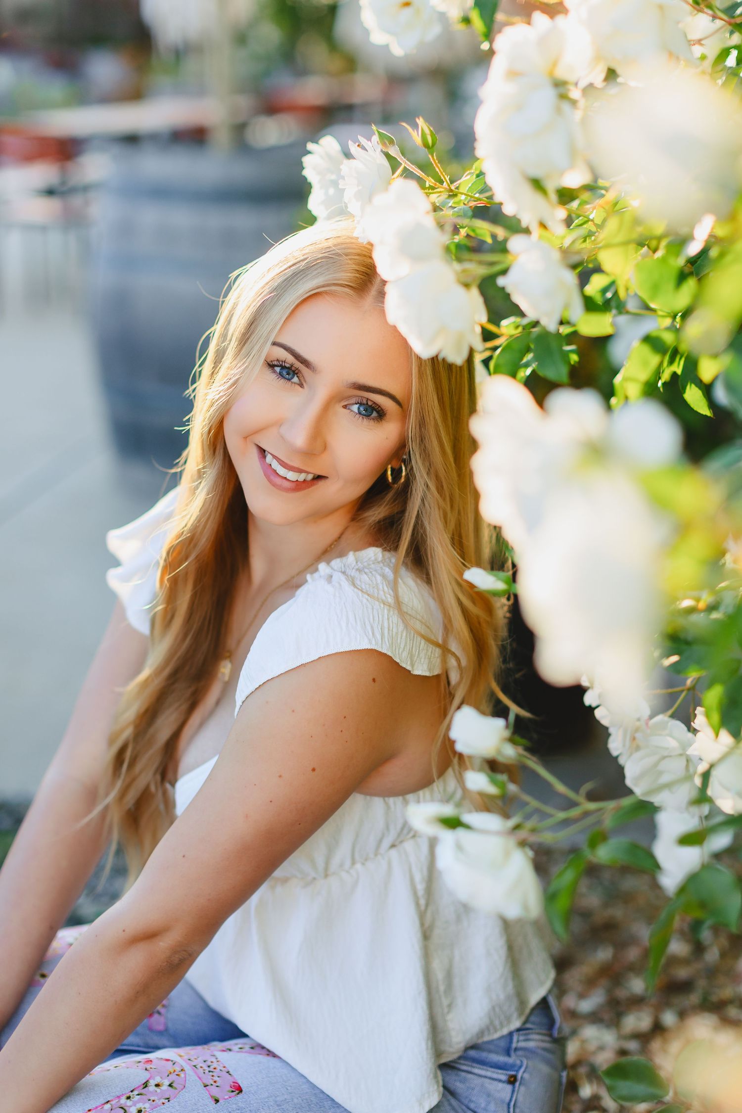 Smiling blonde woman in white top poses near blooming white roses in a sunny outdoor garden setting.