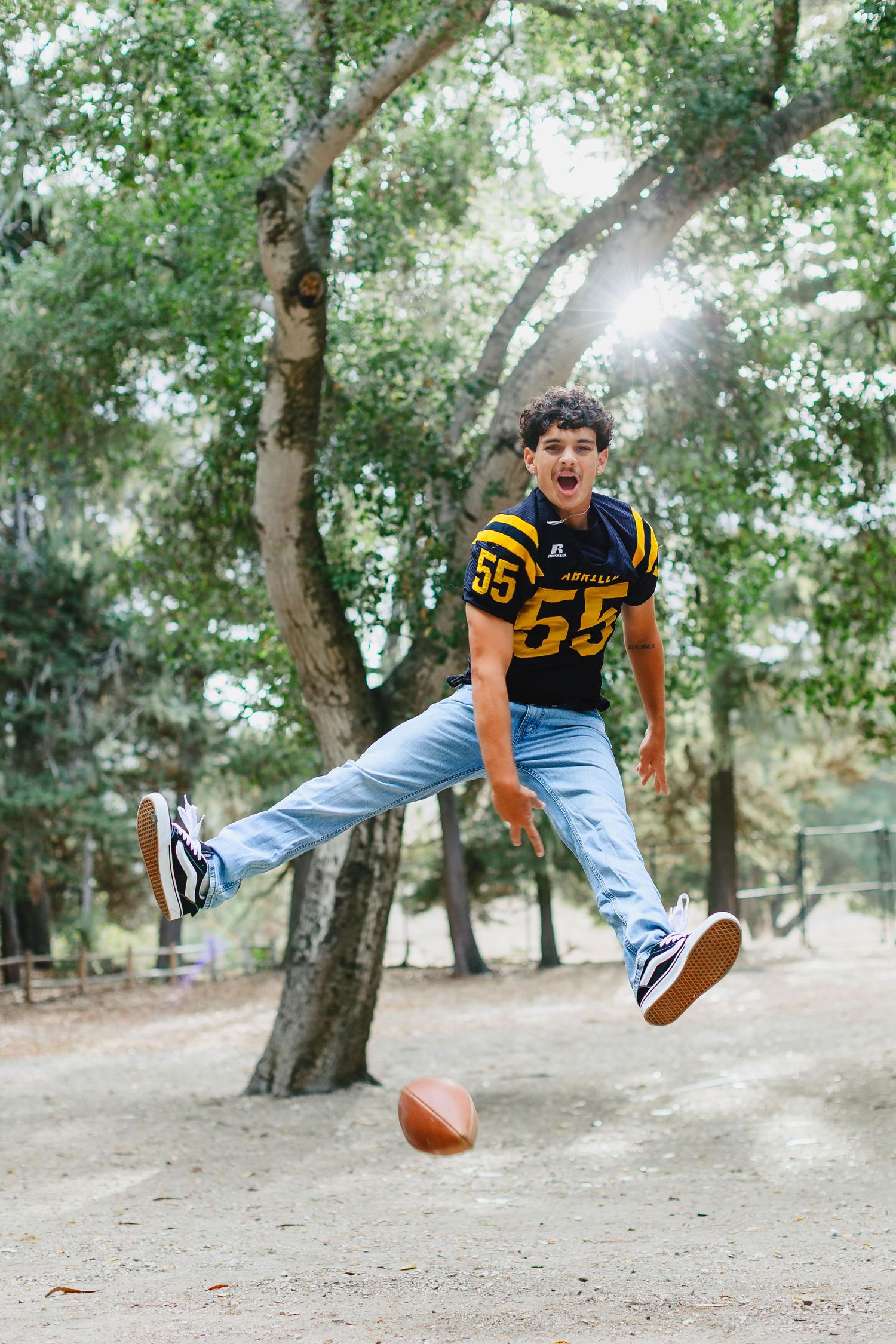 A boy in a football jersey number 55 kicks a football outdoors near trees.