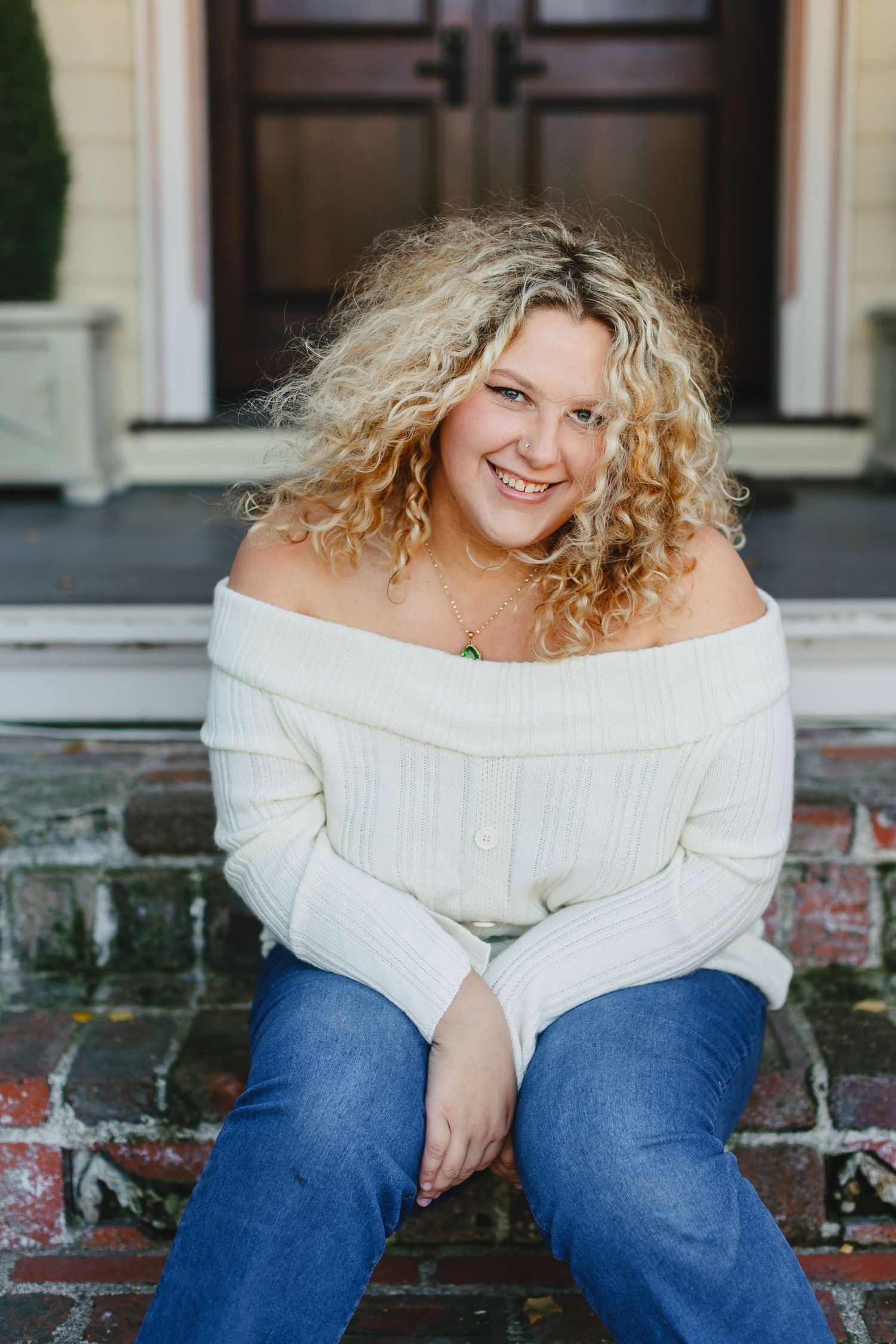 Smiling blonde woman with curly hair sits on brick steps in a white off-shoulder sweater.