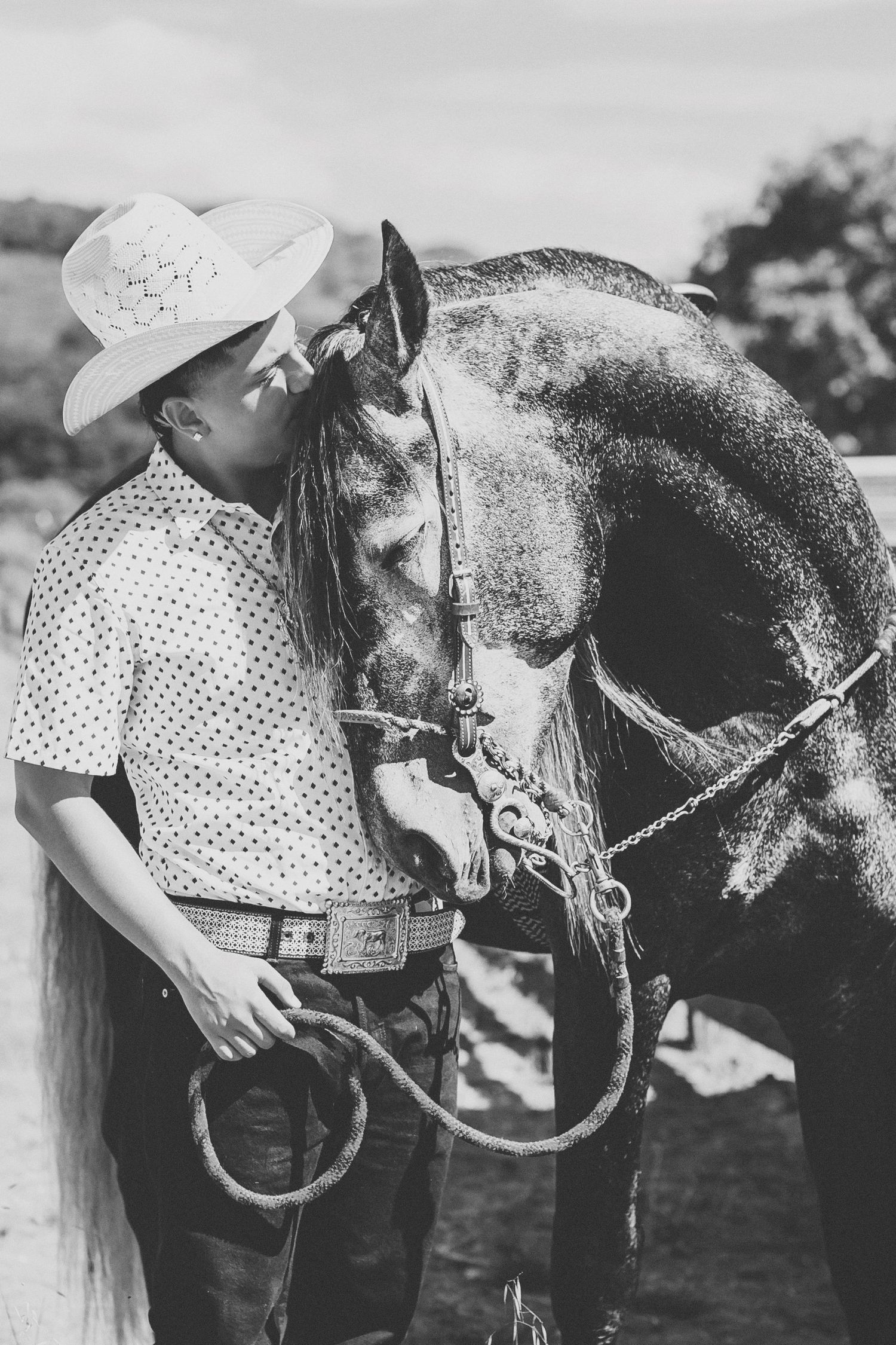 Black and white photo of young person in cowboy hat kissing a dappled horse outdoors.