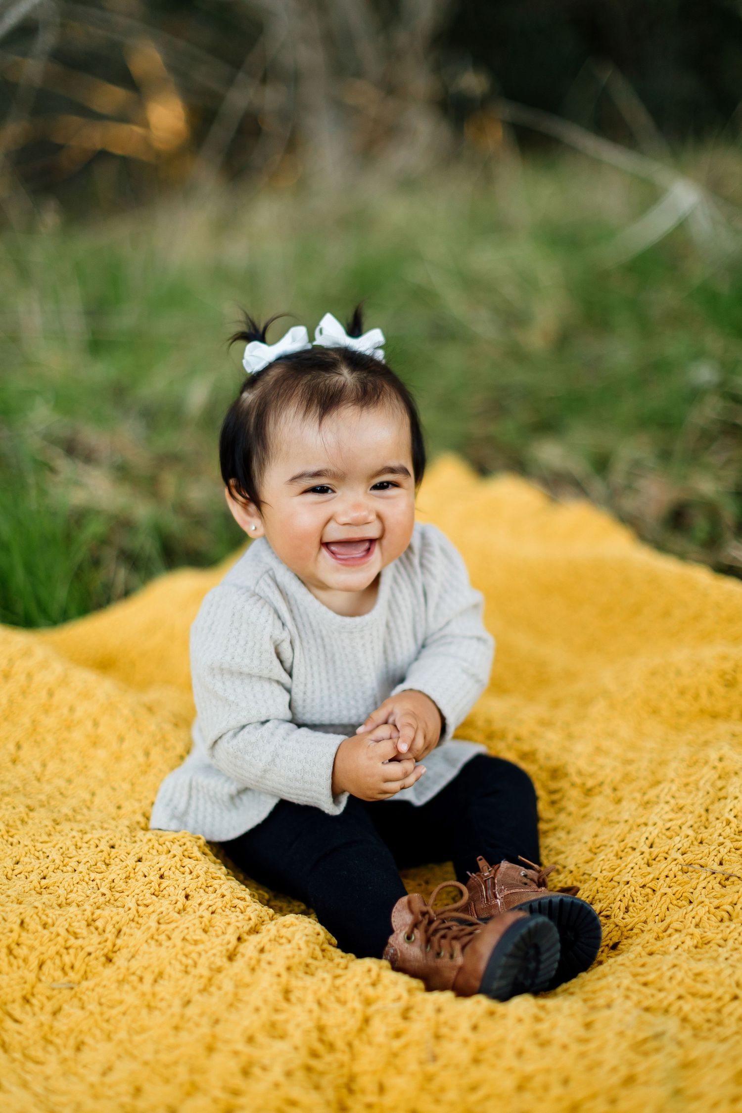 Baby wearing gray sweater and white bow sits on yellow blanket while smiling and laughing.
