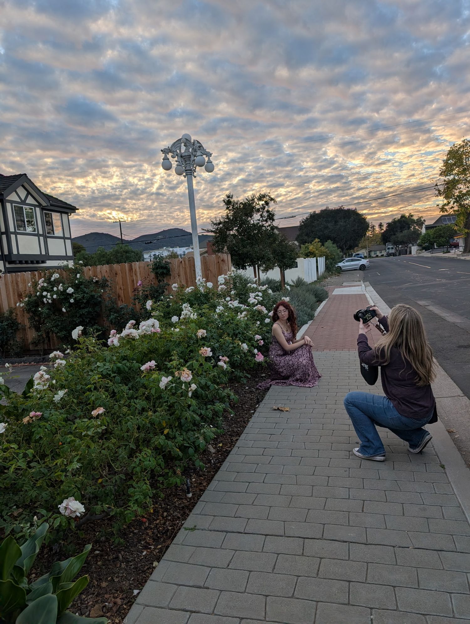 Photographer crouching on brick path capturing woman posing in rose garden at golden hour sunset.