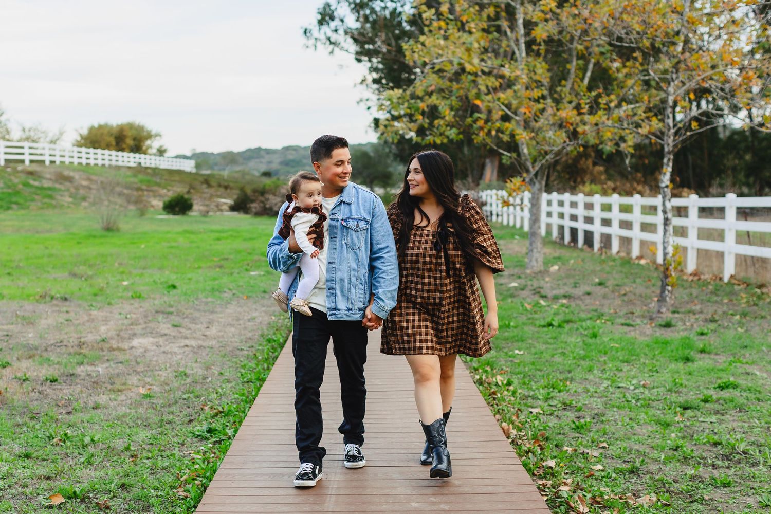 Family of three walking on a wooden path in a park, parents holding toddler, white fence and green grass background.