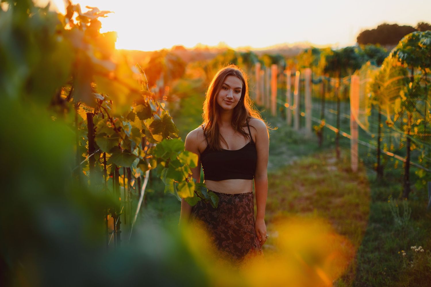 Young woman smiling in a sunlit vineyard at golden hour, surrounded by lush green grapevines.