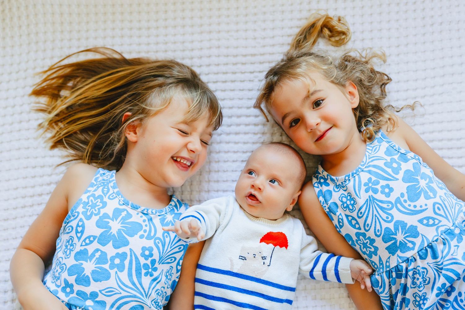 Two young girls in matching blue floral dresses lying down with a newborn baby between them smiling.