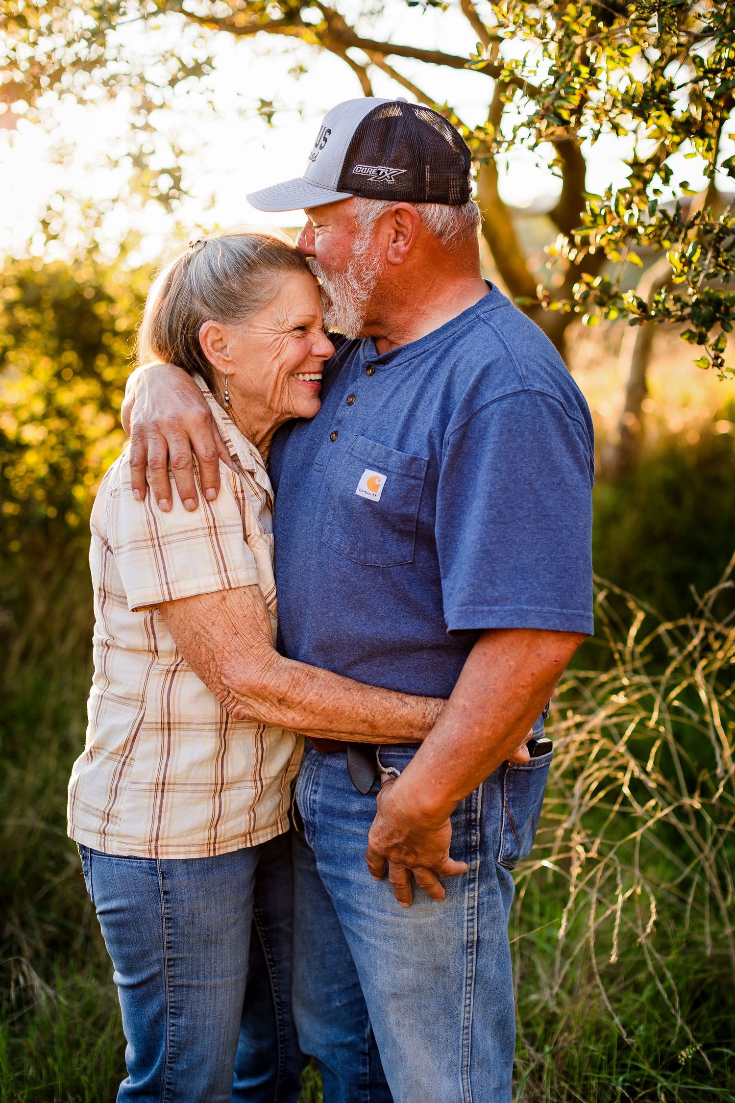 A loving elderly couple embraces outdoors at sunset with golden light filtering through trees in the background.