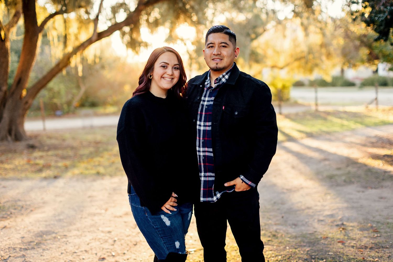 A couple shares a loving embrace on a path during golden hour with sun-lit trees in the background.