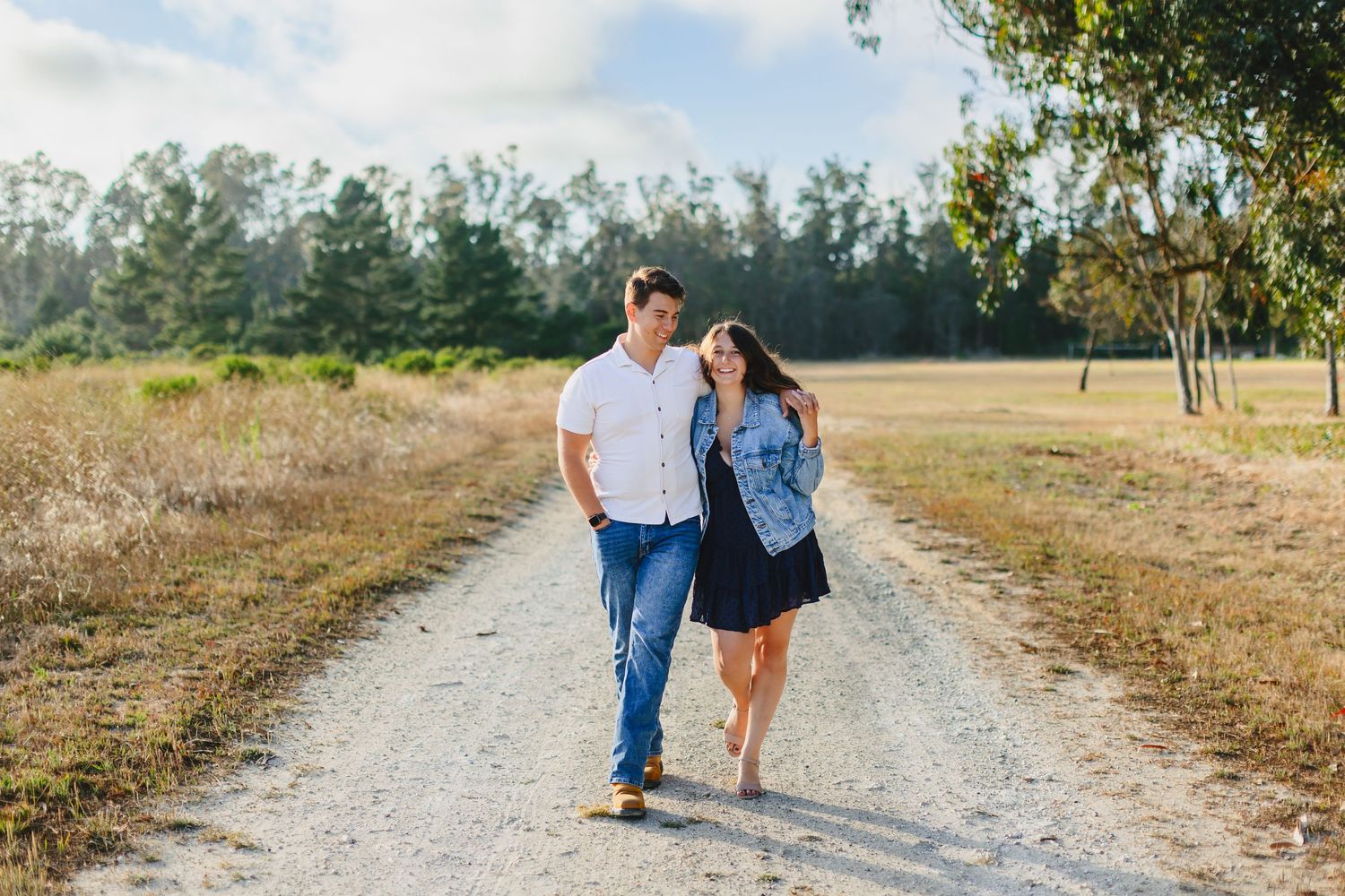 A couple walks together on a sunny dirt path surrounded by golden grass and trees in the distance.