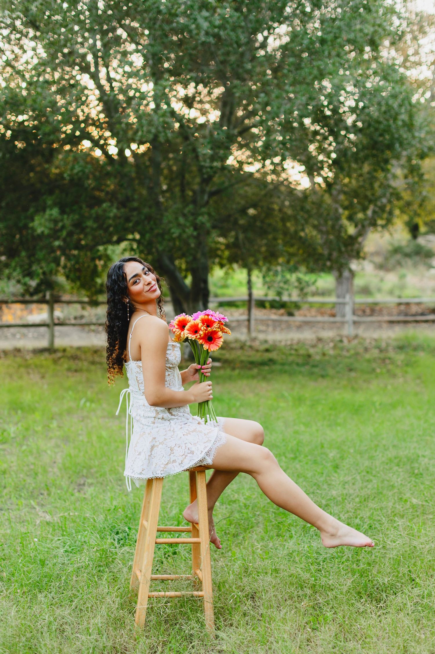 Woman in white dress sits on wooden stool holding colorful flowers in garden setting.
