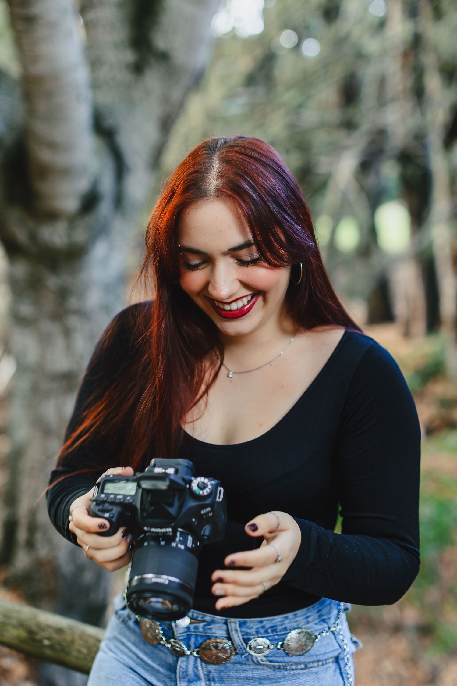 A photographer in a black top and jeans smiles while reviewing photos on their DSLR camera in a wooded setting.