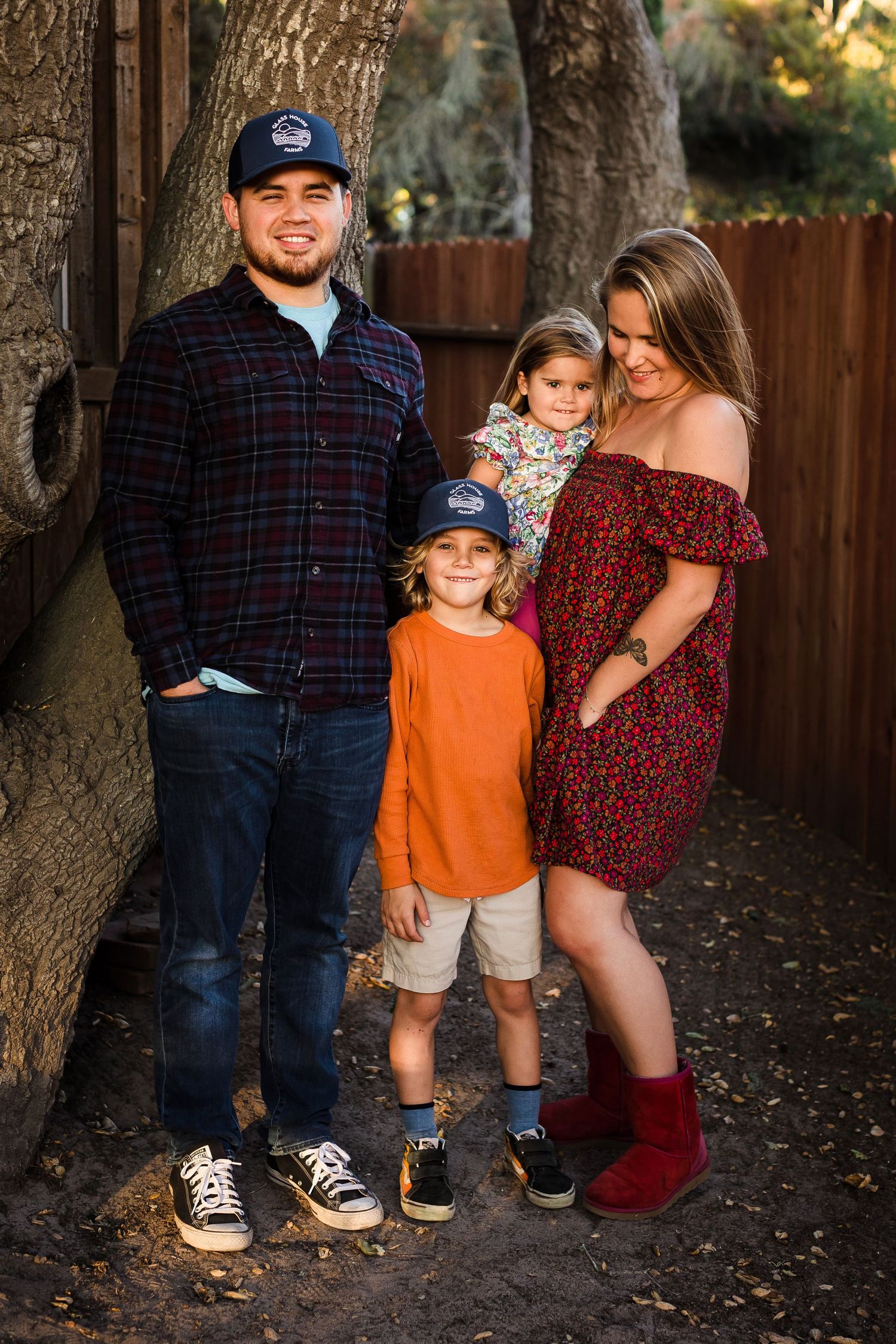 Family group stands close together in autumn setting wearing coordinated casual outfits against wooden fence background.