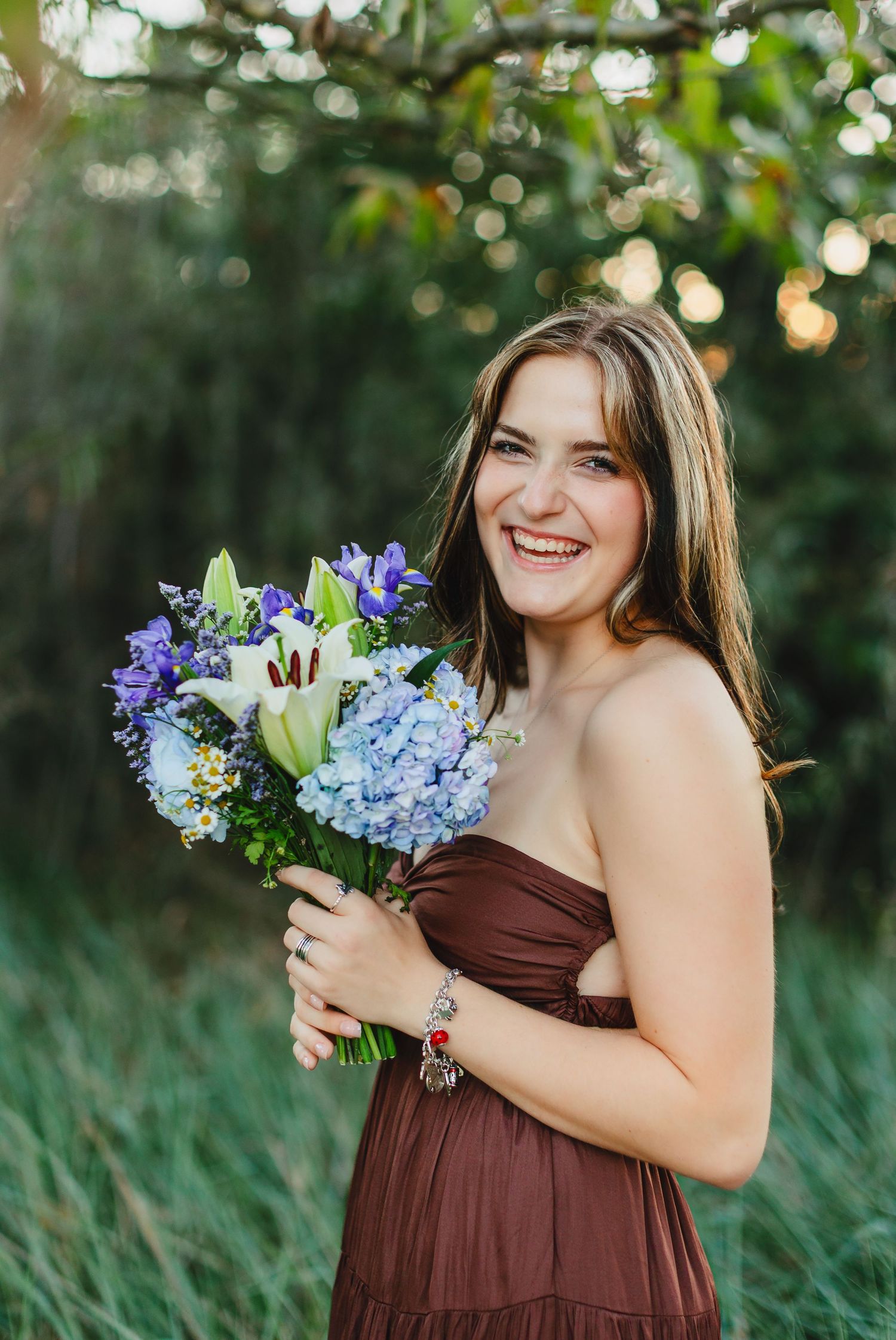Smiling young woman in brown strapless dress holding blue and white flower bouquet outdoors.
