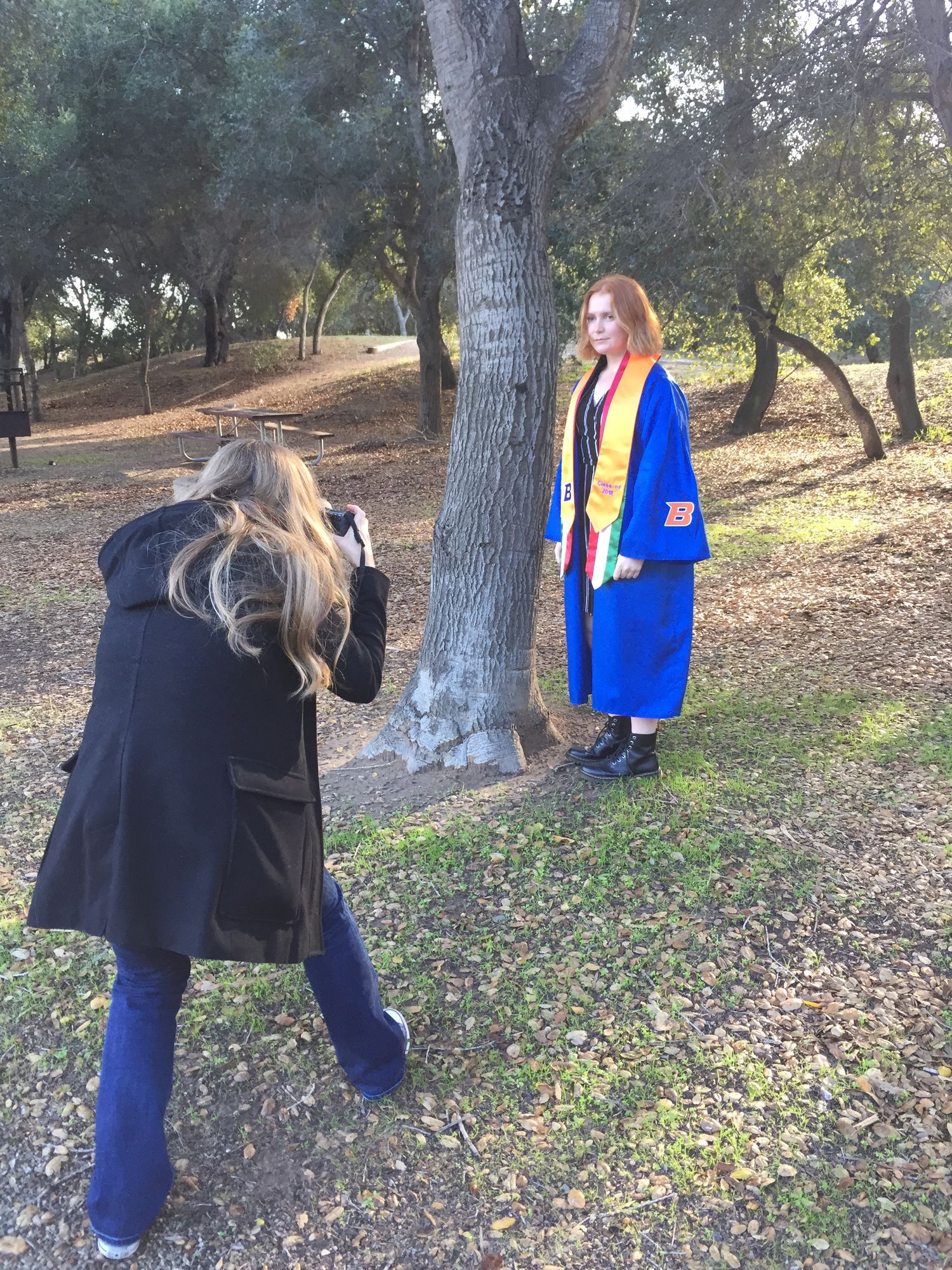 Blonde photographer shooting a redhead student in blue graduation gown near a tree outdoors.