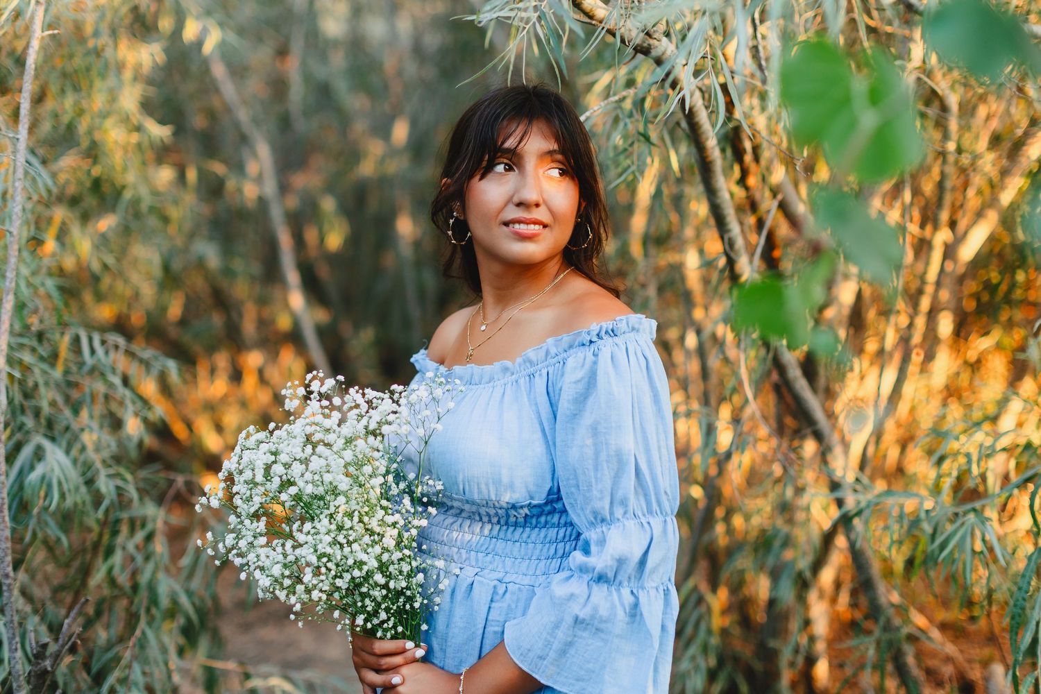 Young woman in blue off-shoulder top holding white wildflowers among green foliage outdoors.