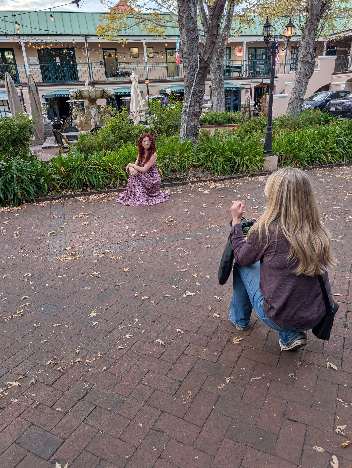 Photographer crouching to capture portrait of woman in purple dress sitting on brick plaza surrounded by greenery.