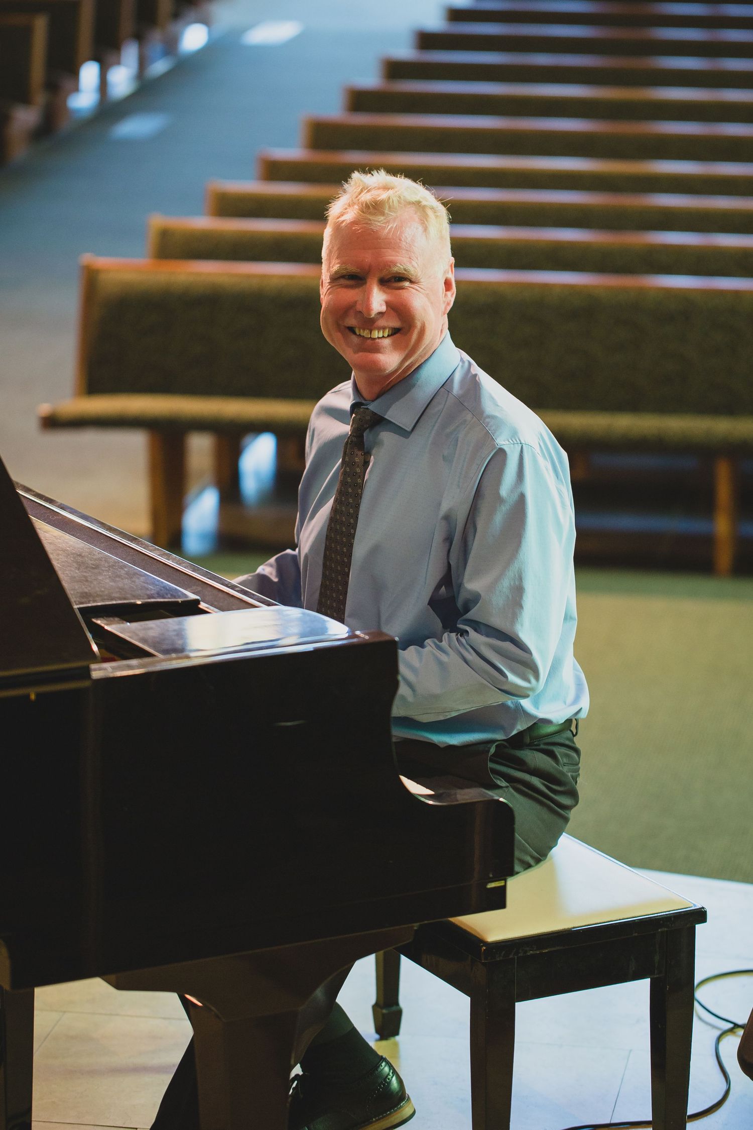 Musician seated at grand piano in performance hall with church pews.