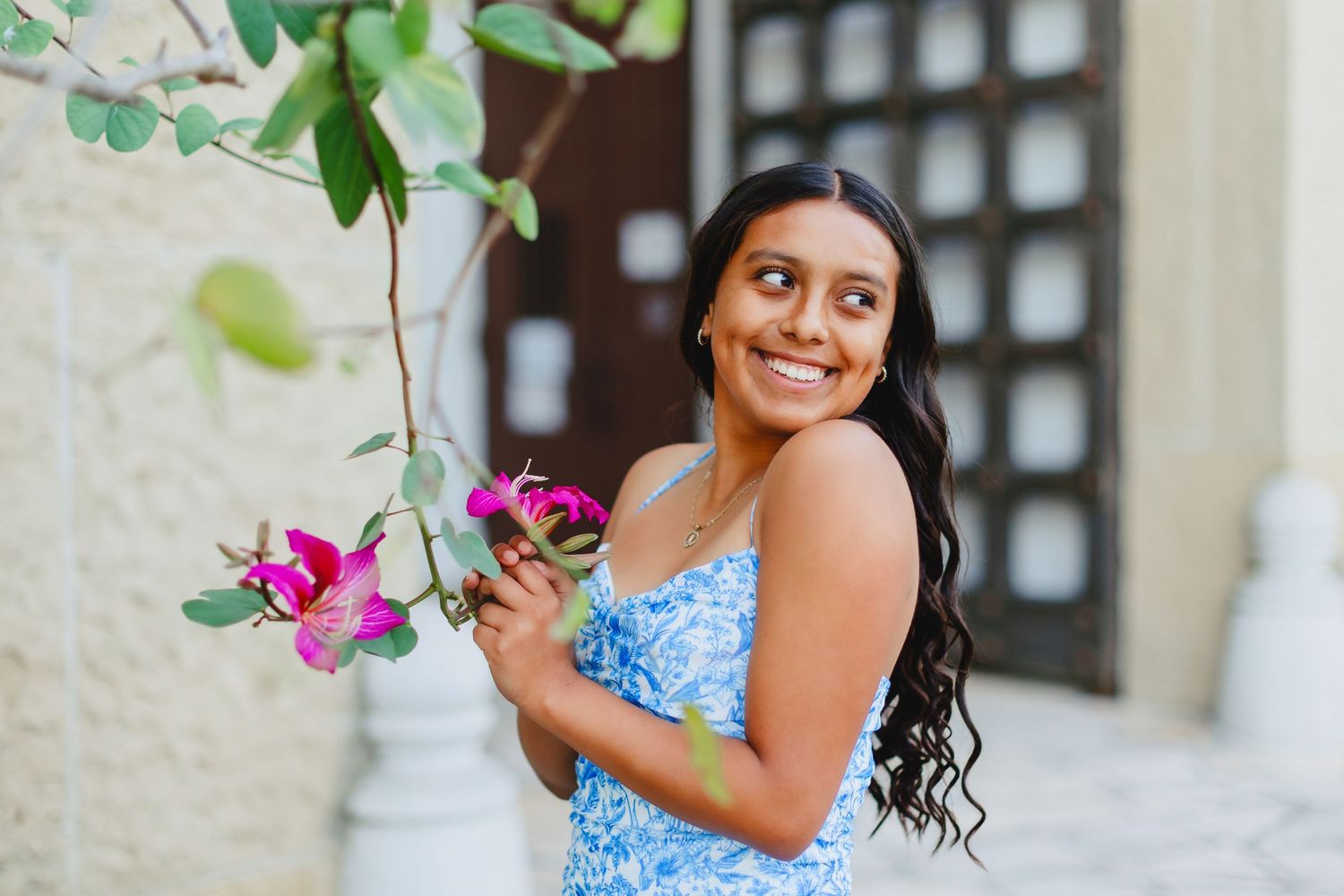 Smiling young woman in blue floral dress holding pink bougainvillea flowers outdoors.