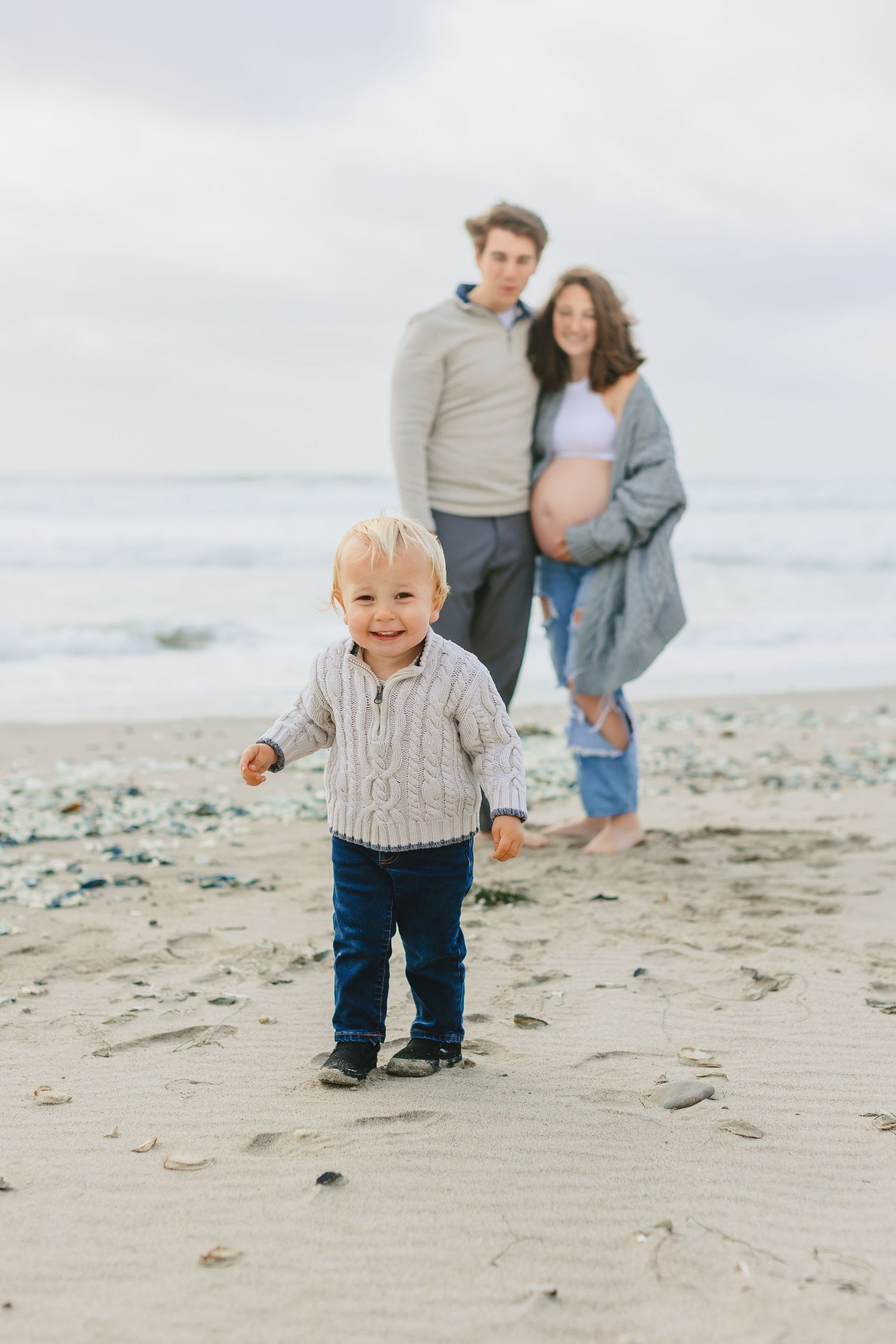A toddler in a knit sweater plays on the beach while others watch in the background.