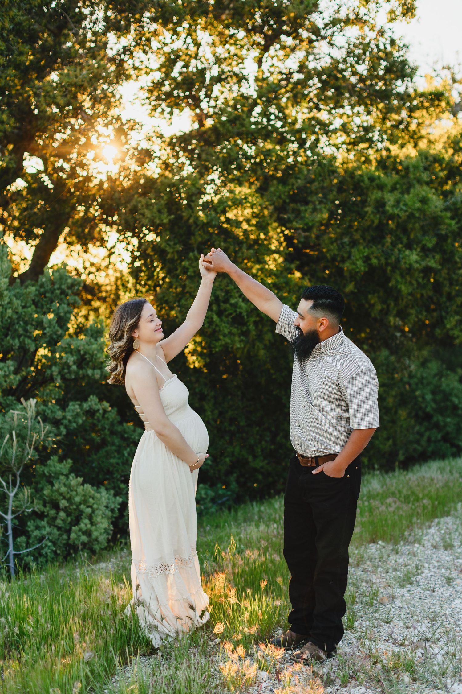 Expecting couple dances joyfully in a sun-dappled meadow during maternity photo session.