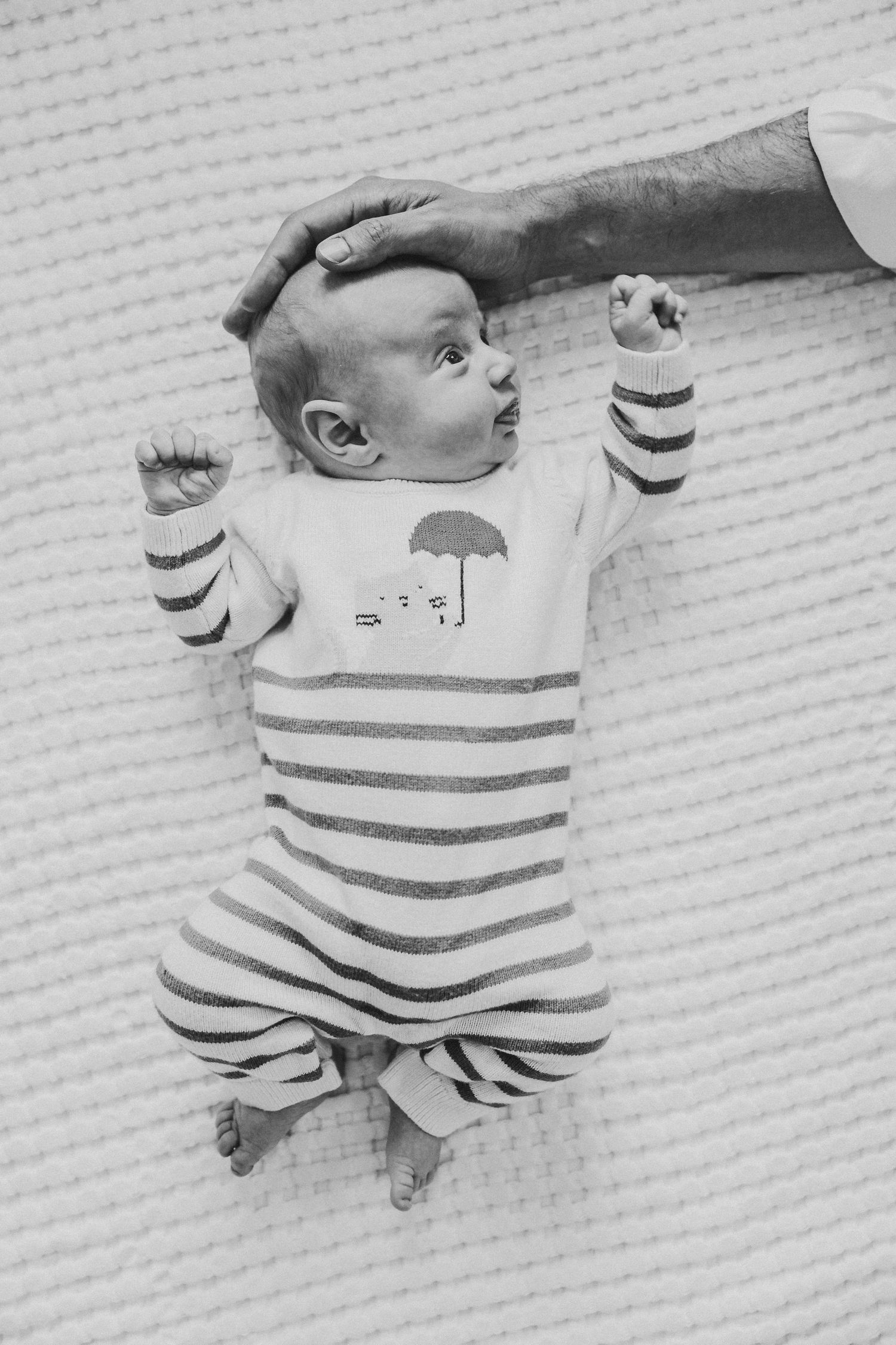 Black and white photo of a newborn baby in striped onesie lying on back with adult hand gently resting on head.