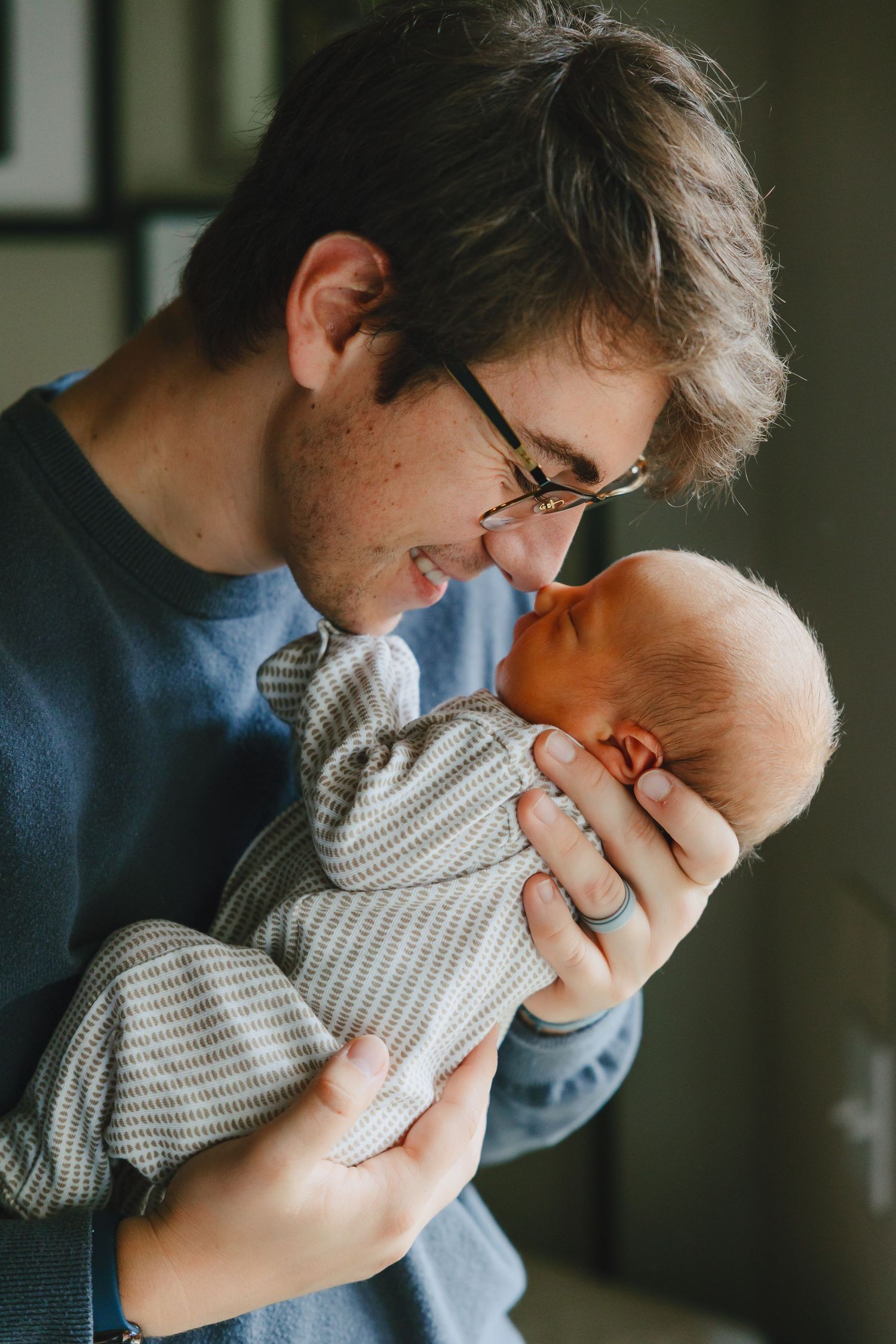 Smiling young father with glasses tenderly holding and nuzzling his newborn baby in a cozy indoor setting.