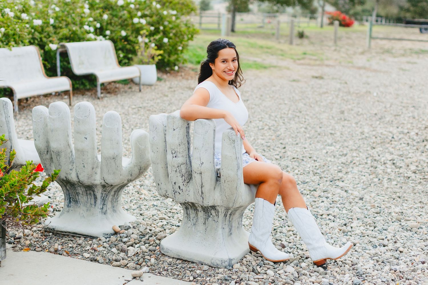 Young woman in white dress and cowboy boots sitting on a concrete hand-shaped bench in an outdoor garden setting.