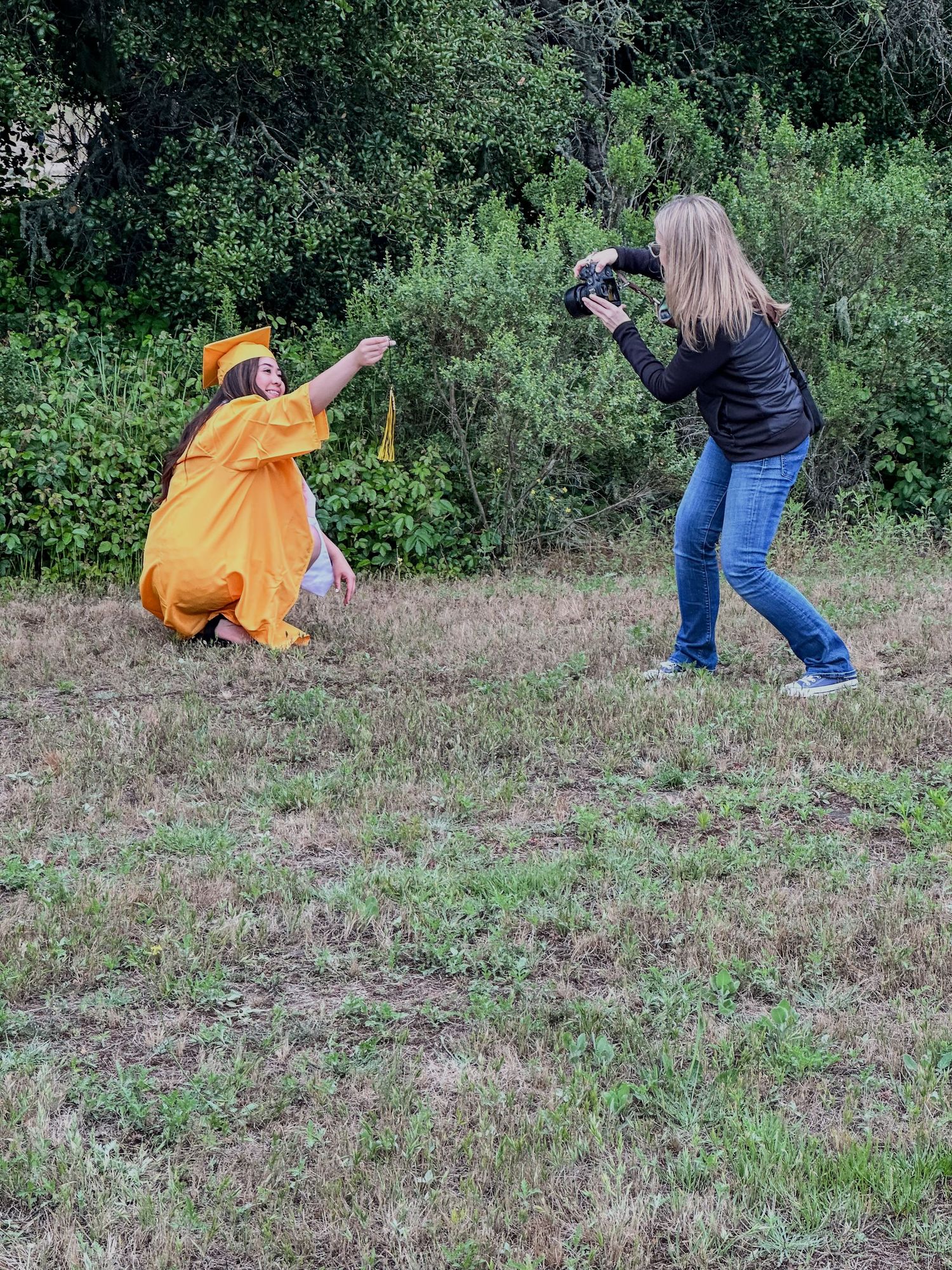 Photographer shooting a graduate in orange gown posing dramatically in a grassy outdoor field.