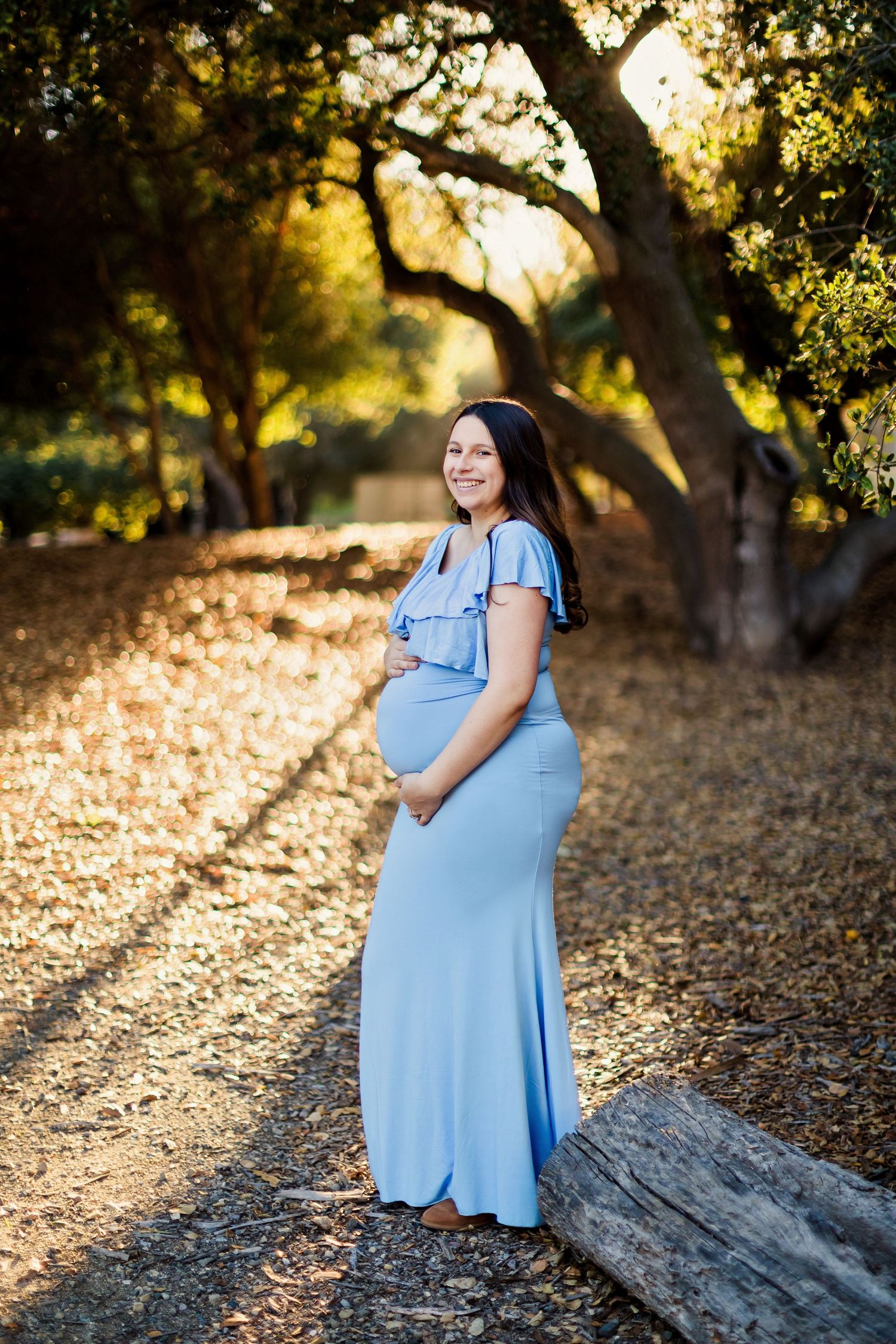 Maternity photoshoot in a golden-lit forest setting with fallen leaves and natural lighting at sunset.