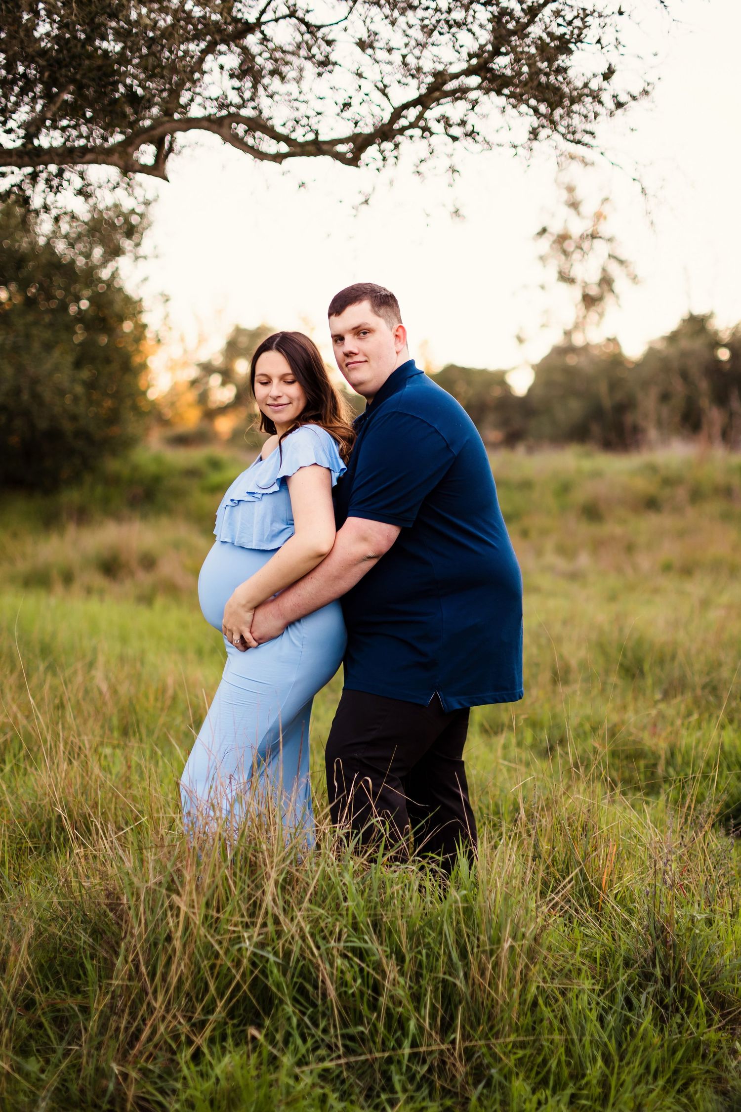 A couple stands together in a grassy field at sunset, with her wearing a light blue dress and him in a navy shirt.