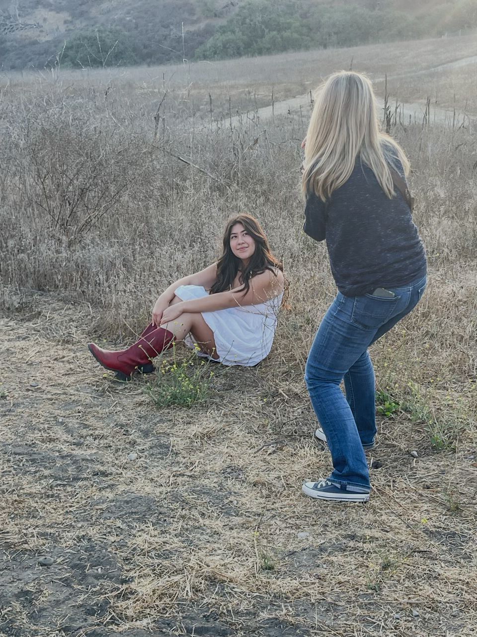 Photographer shooting a young woman in white dress and red boots sitting in a dry grass field.