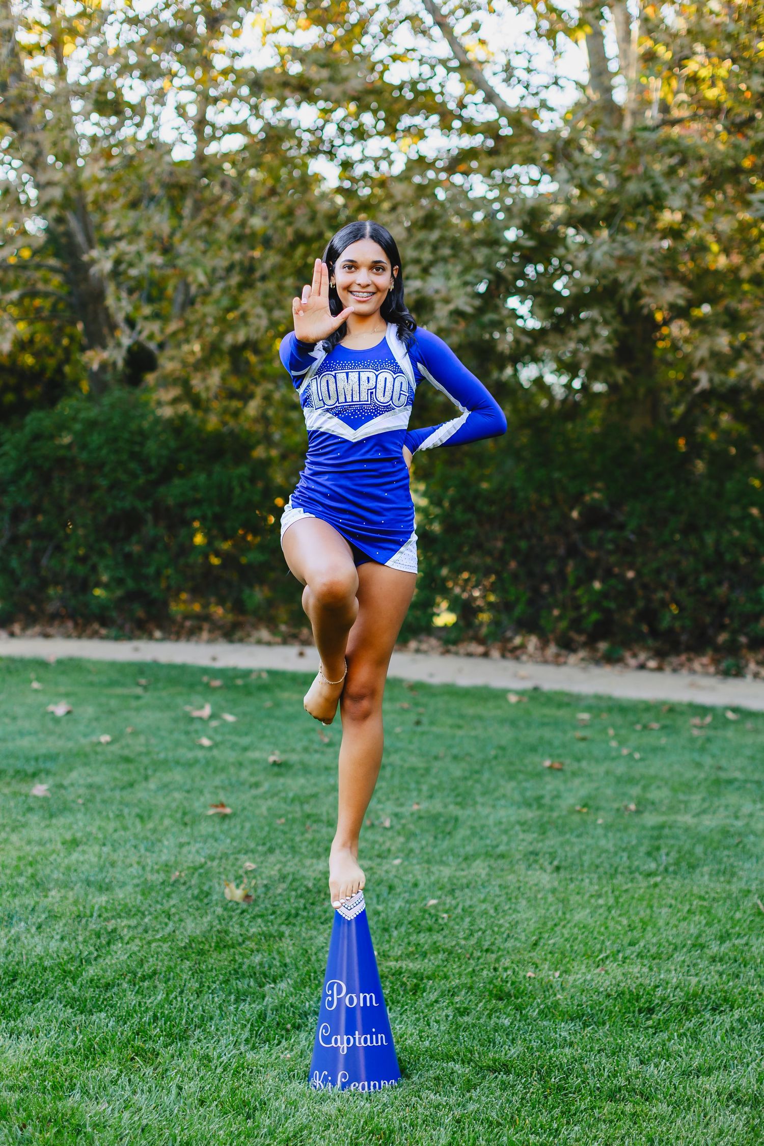 Cheerleader in blue Lompoc uniform balancing on a megaphone outdoors, waving and smiling on green grass.