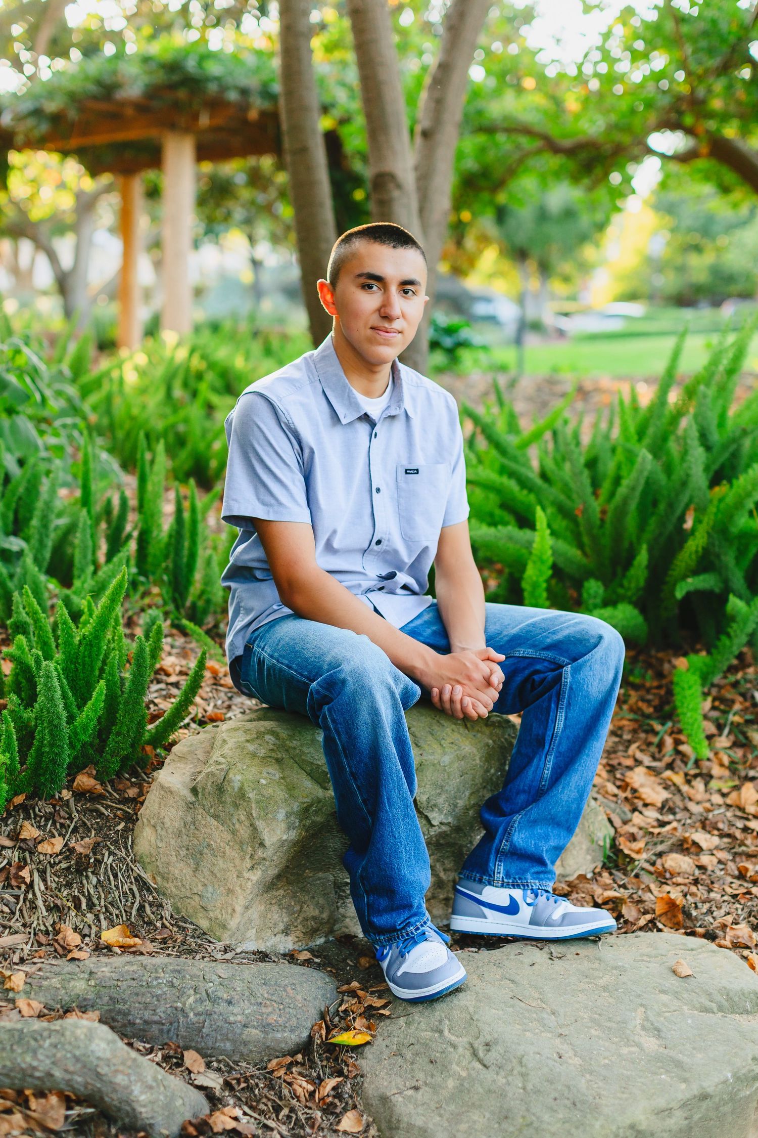 Young man in blue shirt and jeans sitting on a rock in a lush green park setting.